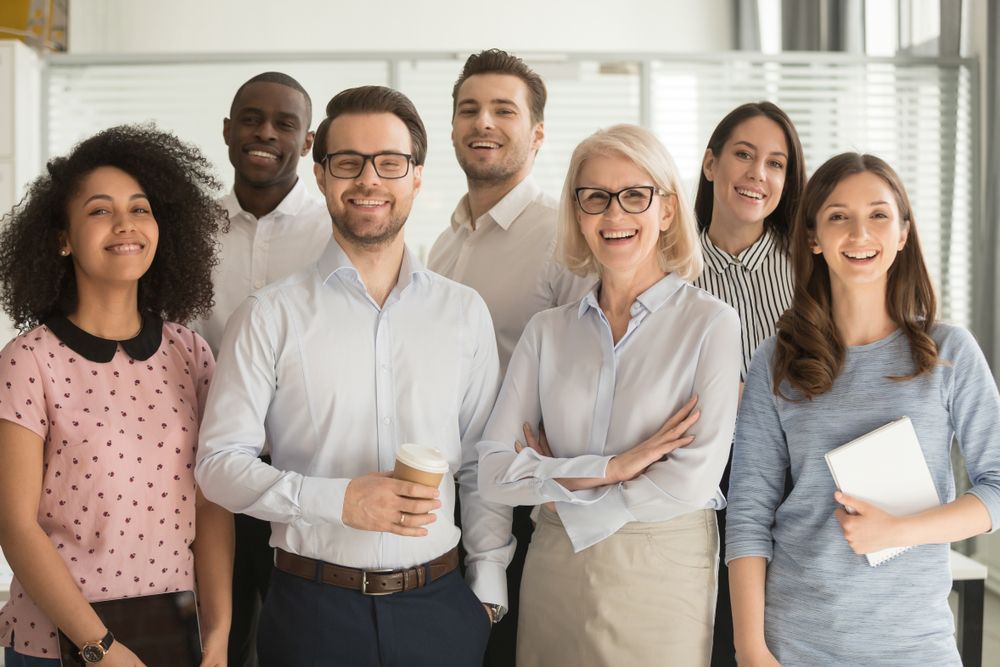 Six smiling coworkers standing together in a bright office, holding coffee, documents, and notebooks.