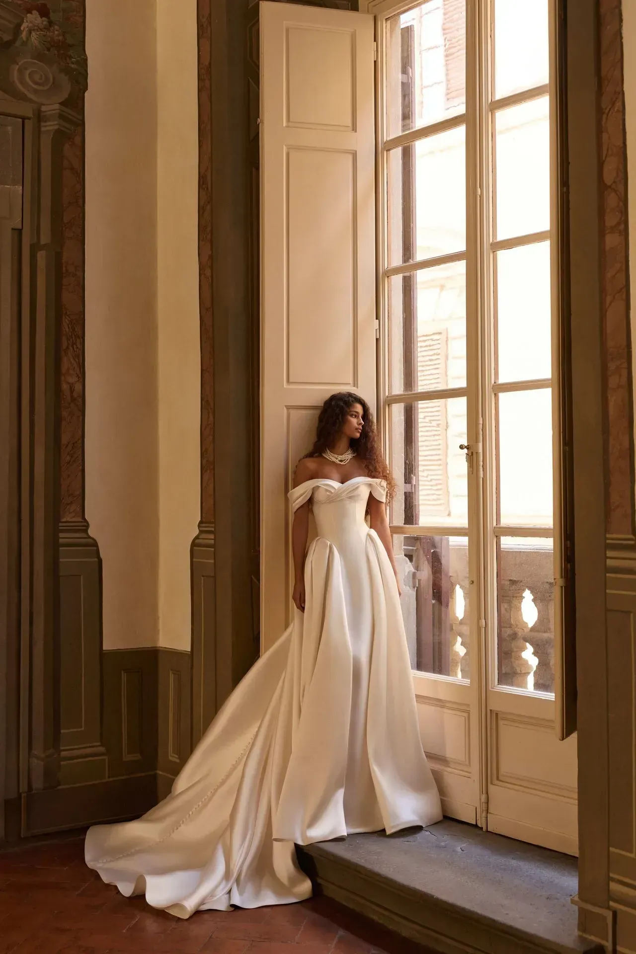 A-line mikado gown with a structured corset, a long skirt with a sweeping train, hidden pockets, and a row of decorative buttons down the back. Draped off-the-shoulder straps add softness to the clean silhouette.
Best complemented with the long-sleeved lace Sylvielle bolero for a vintage-inspired finish.