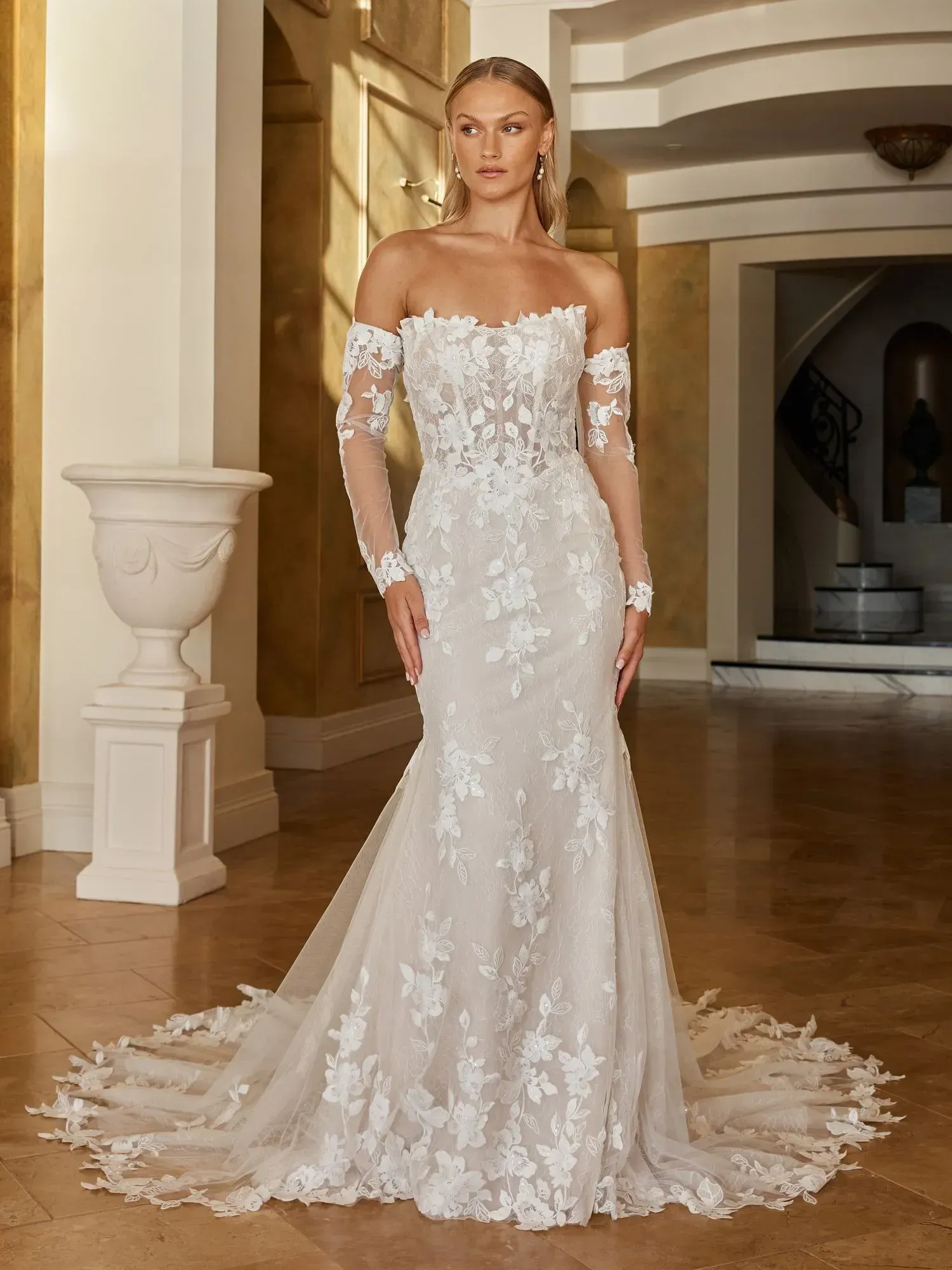 With all the signature features that our Madi brides truly love, our Ohana wedding dress is a lace-forward, fit and flare design that is sexy and glamorous. This gown flatters the figure in all the best ways with its structured fit, while softened with delicate Chantilly lace and subtle shimmer.
Ohana’s bodice is a modern, strapless shape with a straight neckline coming to soft cat-eye peaks. Trimmed with an organic lace edge to soften the modern lines. As a sheer illusion bodice, corset boning construction can be seen peeking through the lace, giving the gown it’s snatched silhouette. Further highlighted by the Basque waistline, drawing in the figure.
For a second look, Ohana can transform with matching detachable lace and tulle long sleeves. Sitting off-the-shoulder, with motifs around the cuffs and running up the sides of the arm. Adding extra coverage and hitting trends.