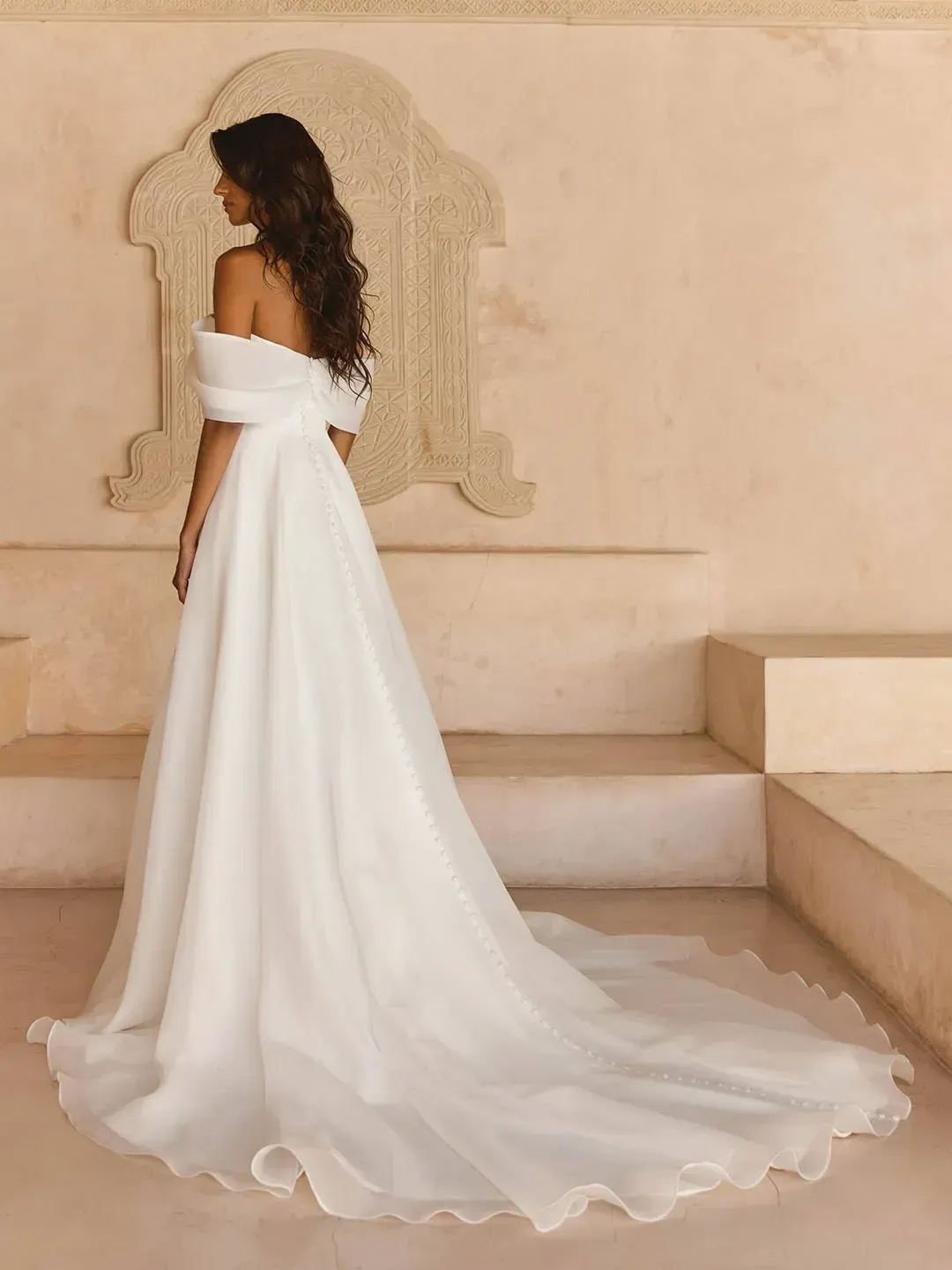 LISSY
Where romance and simplicity meet, you will find our Lissy wedding dress from our Solara collection.  
Whilst, Lissy is not adorned in lace or sparkle she is far from plain, made from our signature organza fabric which is beautifully lightweight and creates an ethereal soft look. 
Matching the softness of the fabric is the equally soft off-the-shoulder neckline featuring a draped organza cuff giving the most delicate texture.  
Her asymmetric pleated organza bodice creates a snatched shape in combination with her modern Basque waistline. She is constructed with our signature boning, and additional internal banded support for total security, comfort, and the most flattering fit. 
Designed to have the frothiest lightweight A-line skirt, Lissy delivers soft drama and even features side seam pockets.  
Around the back, she sits higher for optimal support across all sizes, with the bodice featuring the same asymmetric pleated detail.  
Organza-covered buttons complete the look by running all the way down her illusion organza train for the perfect effortless and romantic look.  