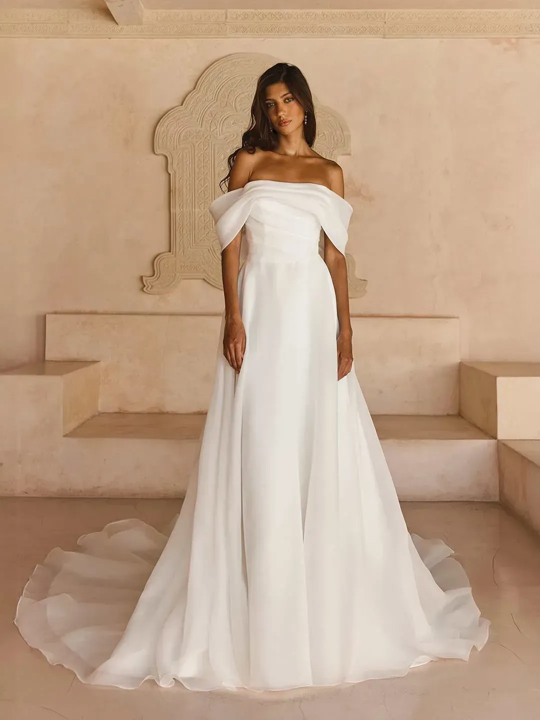 LISSY
Where romance and simplicity meet, you will find our Lissy wedding dress from our Solara collection.  
Whilst, Lissy is not adorned in lace or sparkle she is far from plain, made from our signature organza fabric which is beautifully lightweight and creates an ethereal soft look. 
Matching the softness of the fabric is the equally soft off-the-shoulder neckline featuring a draped organza cuff giving the most delicate texture.  
Her asymmetric pleated organza bodice creates a snatched shape in combination with her modern Basque waistline. She is constructed with our signature boning, and additional internal banded support for total security, comfort, and the most flattering fit. 
Designed to have the frothiest lightweight A-line skirt, Lissy delivers soft drama and even features side seam pockets.  
Around the back, she sits higher for optimal support across all sizes, with the bodice featuring the same asymmetric pleated detail.  
Organza-covered buttons complete the look by running all the way down her illusion organza train for the perfect effortless and romantic look.  