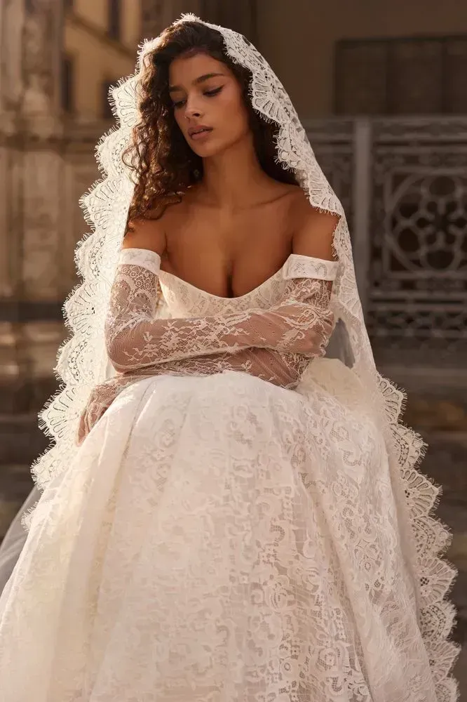 Chantilly lace princess gown with a corset bodice, detachable beaded trim at the bust that you can remove if you want a cleaner, more minimal look, and detachable off-shoulder sleeves for an easy second-look switch. Finished with a lush skirt and decorative buttons down the back.
Style it with the Elismera veil and Odyssia gloves for a romantic, artfully composed bridal look.
