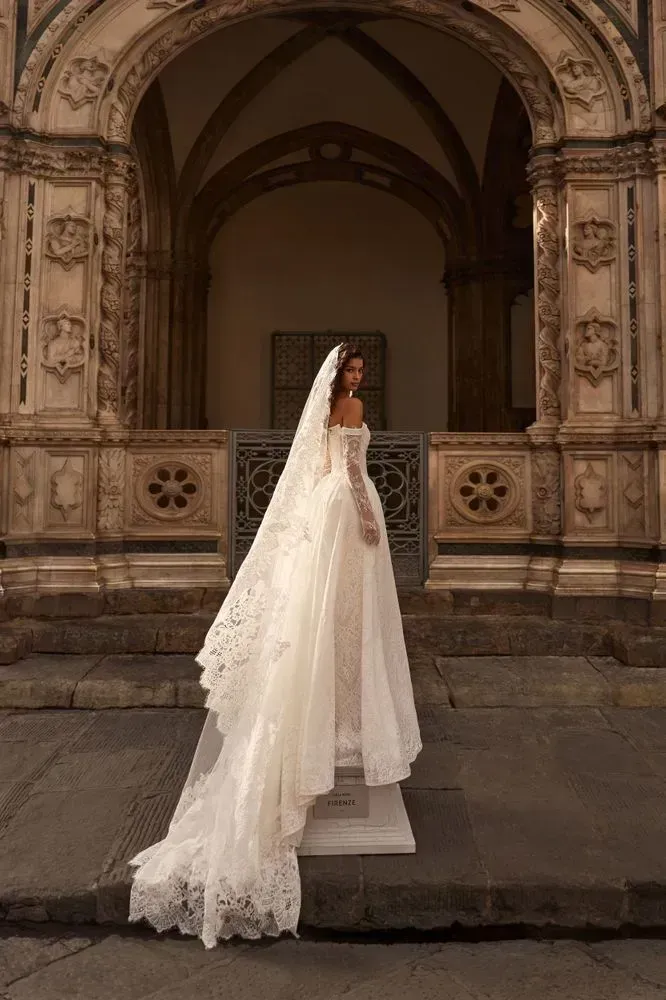 Chantilly lace princess gown with a corset bodice, detachable beaded trim at the bust that you can remove if you want a cleaner, more minimal look, and detachable off-shoulder sleeves for an easy second-look switch. Finished with a lush skirt and decorative buttons down the back.
Style it with the Elismera veil and Odyssia gloves for a romantic, artfully composed bridal look.