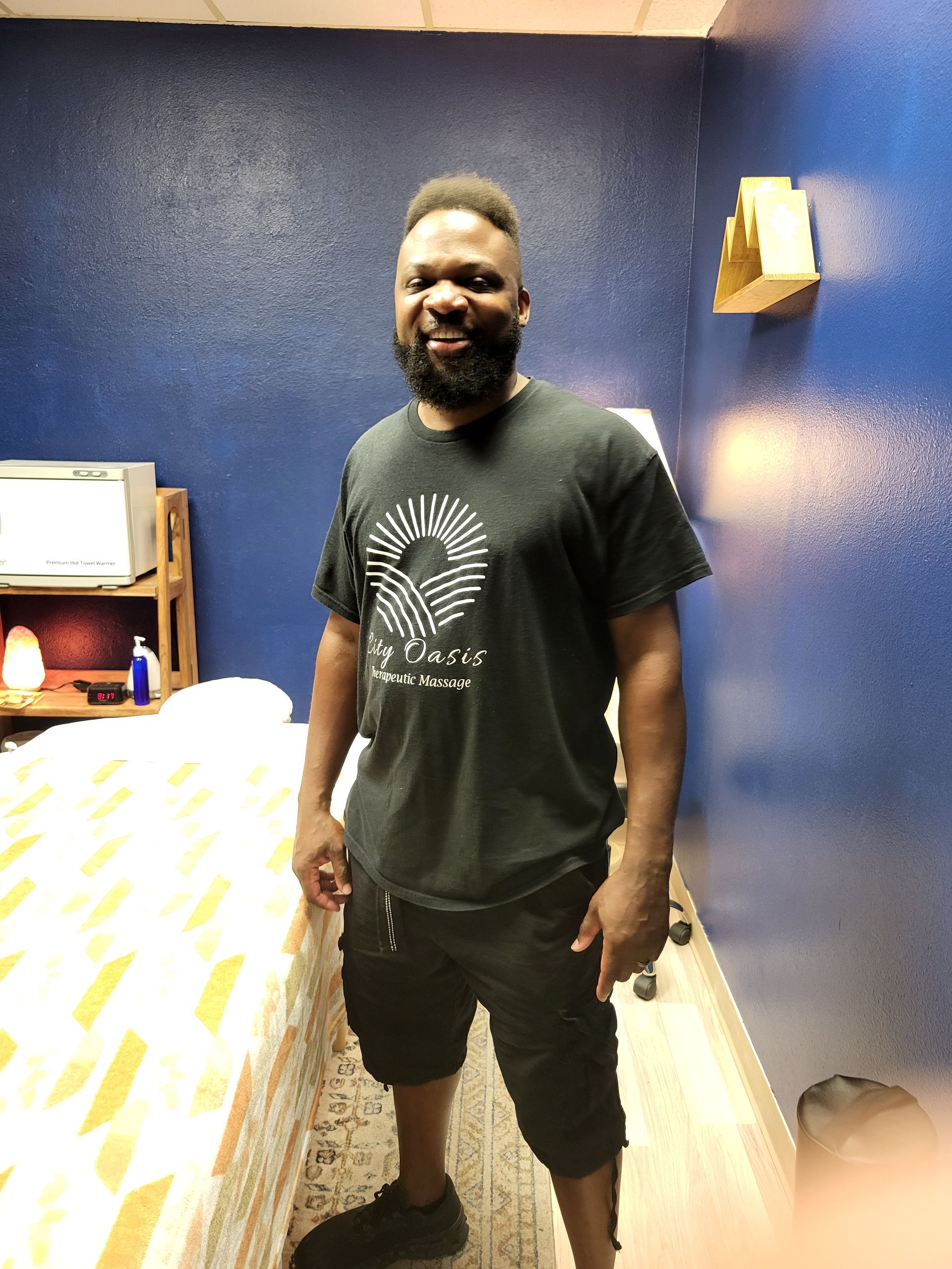 Smiling man standing in a room with blue walls and warm lighting, wearing a black T-shirt and shorts