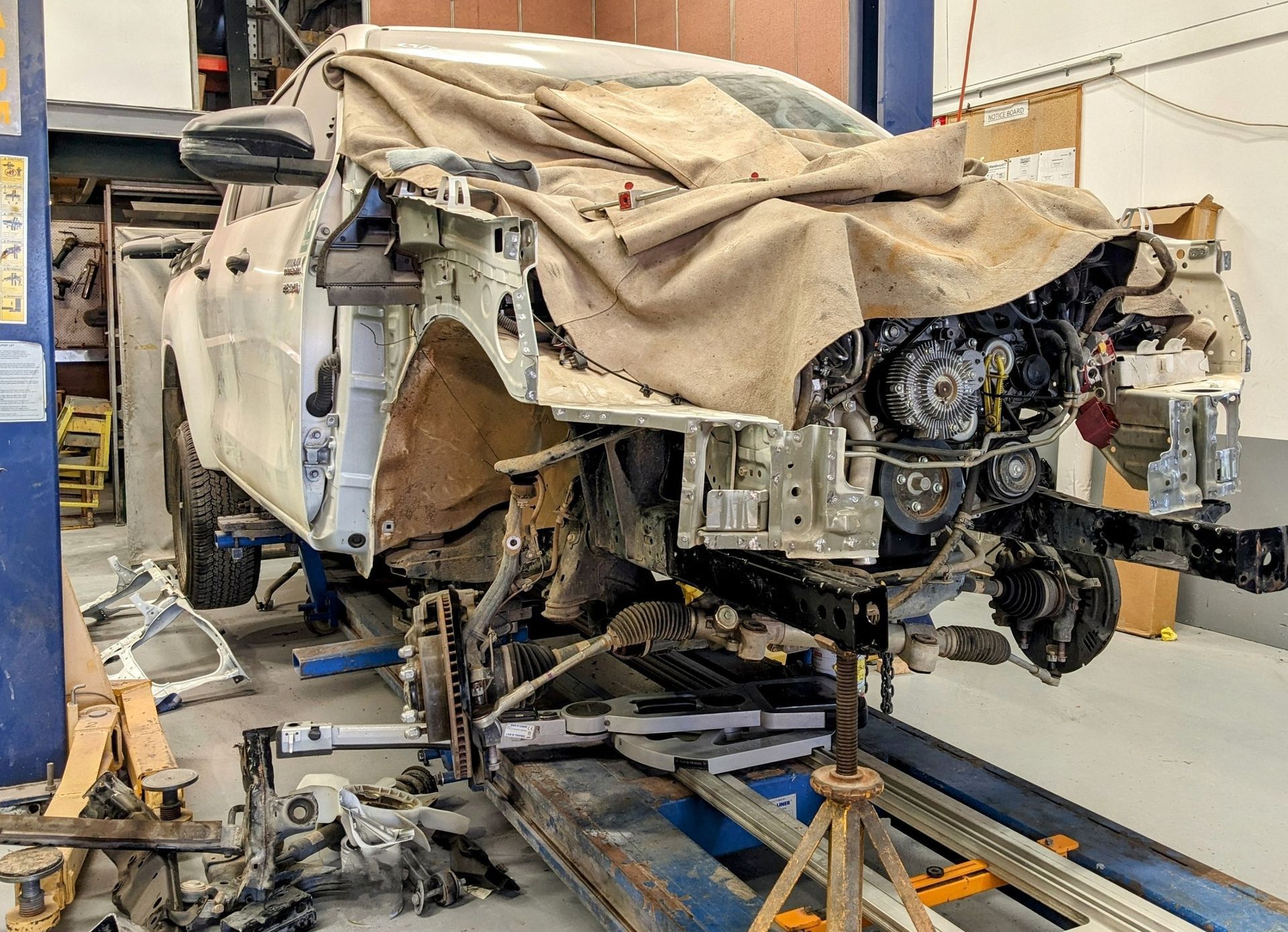A disassembled white vehicle with its front end exposed, suspended on a hydraulic lift inside an auto repair workshop — Supalook Smash Repairs In Port Macquarie, NSW