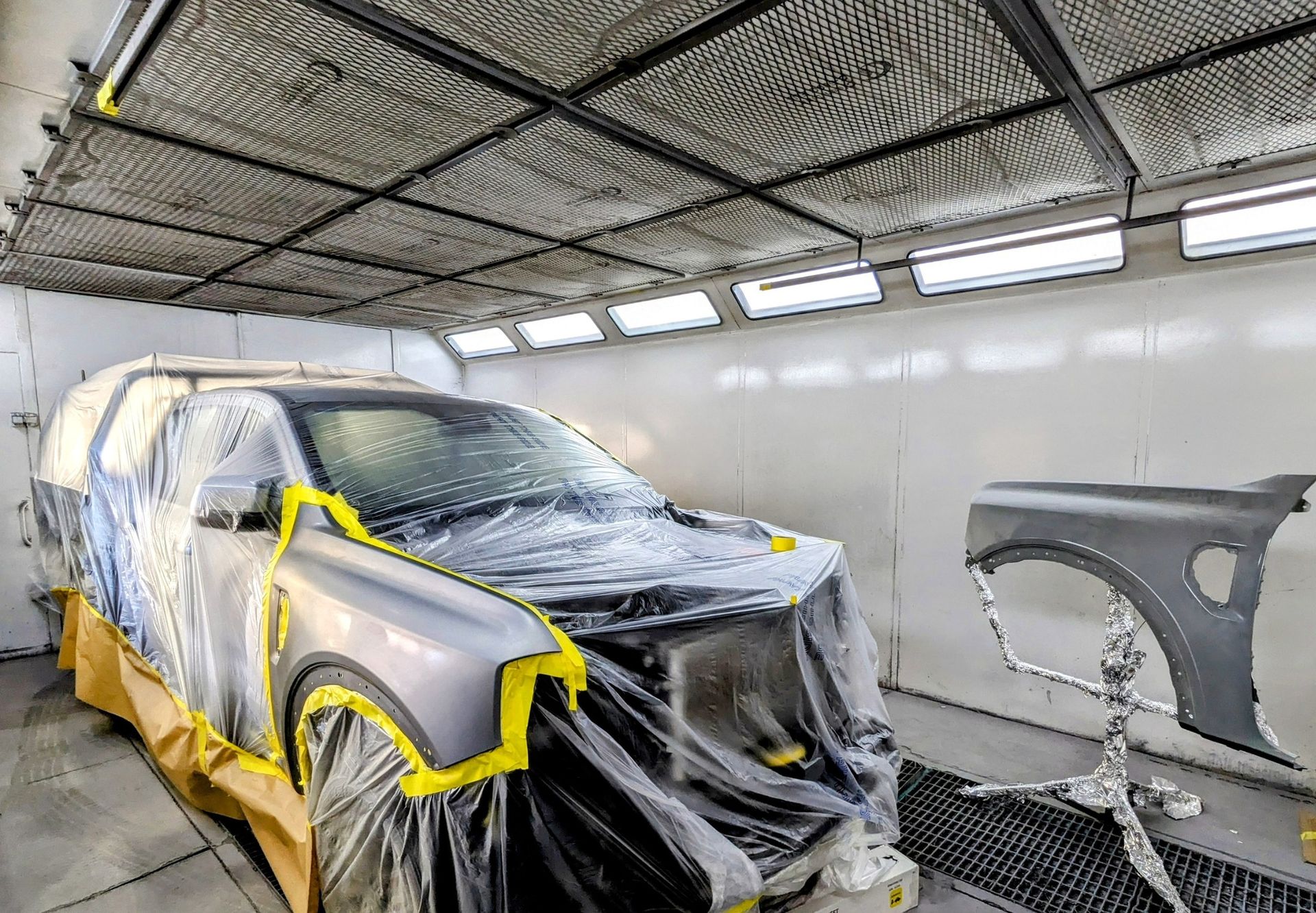 A silver pickup truck masked with tape and plastic sheeting inside an industrial paint booth with a detached fender — Supalook Smash Repairs In Port Macquarie, NSW
