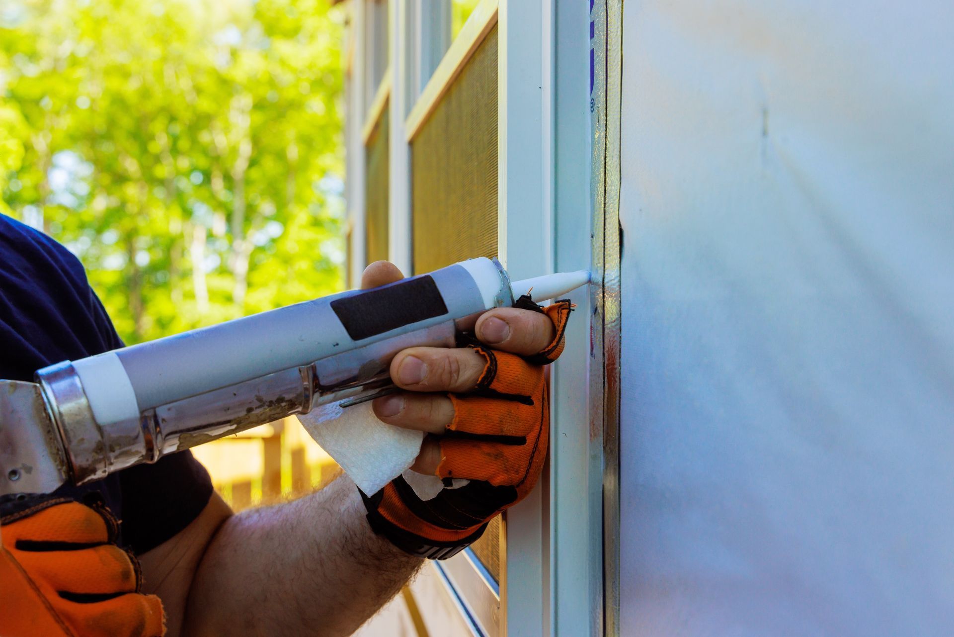Gloved hand applying caulk with a caulking gun along a window frame outdoors
