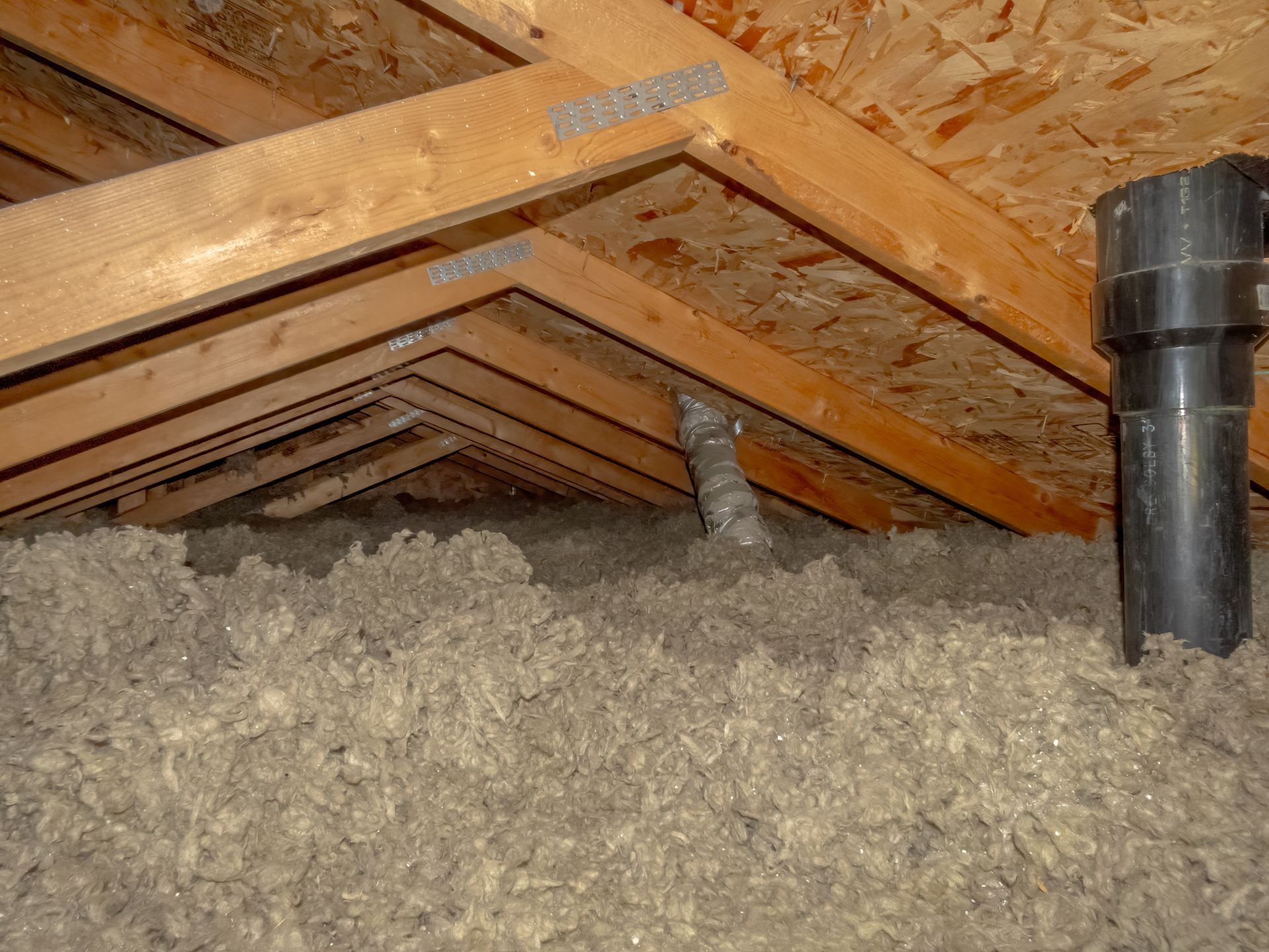 Attic with exposed wooden rafters, insulation, and a black vent pipe in the foreground