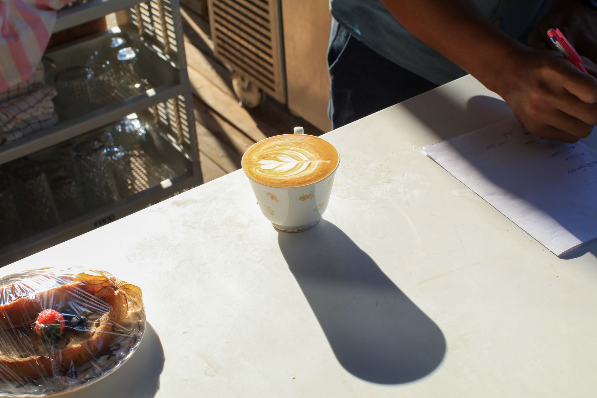 Coffee cup and pastry on a sunny cafe table, with a person's arm writing nearby.