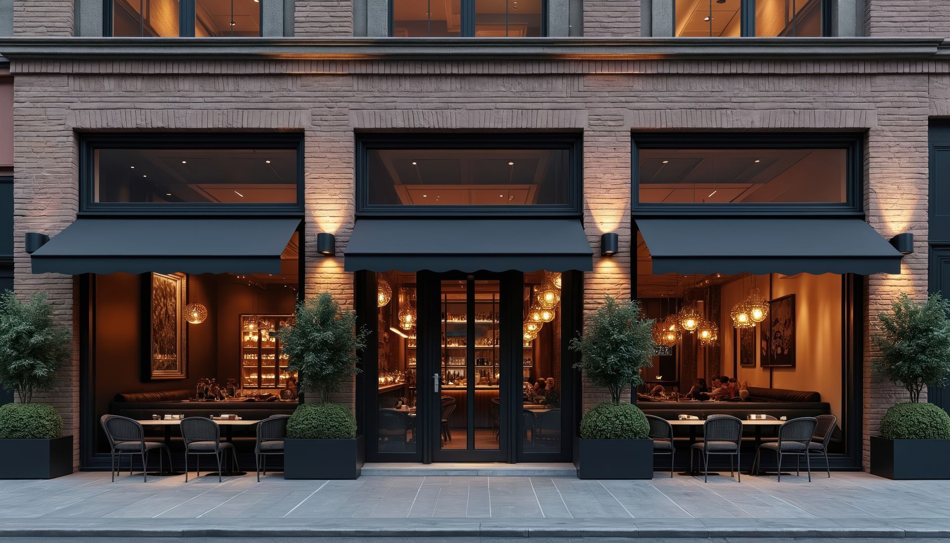 Street-level view of a brick café with black awnings, warm interior lighting, and outdoor tables