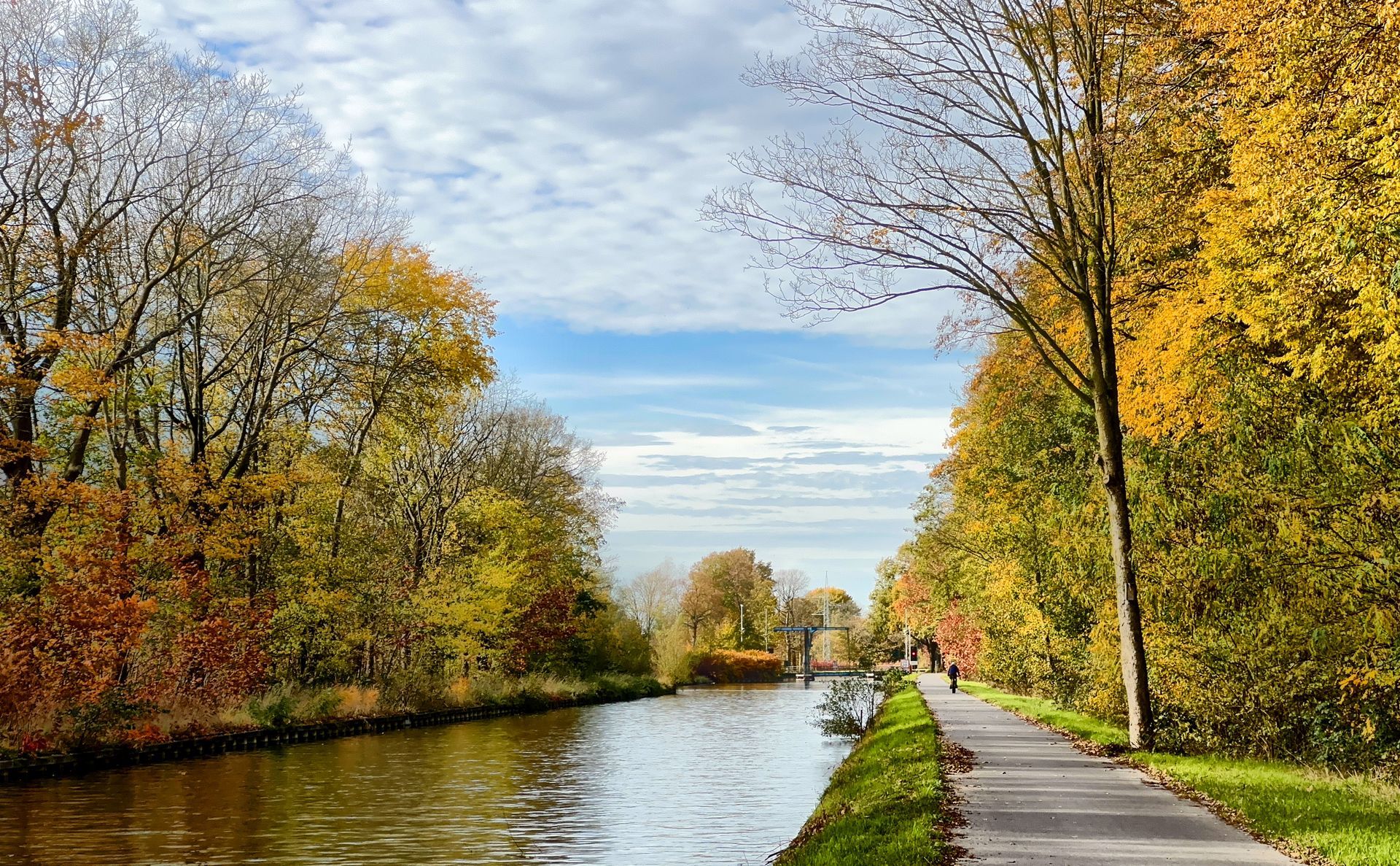 Canal lined with autumn trees beside a walkway under a cloudy sky