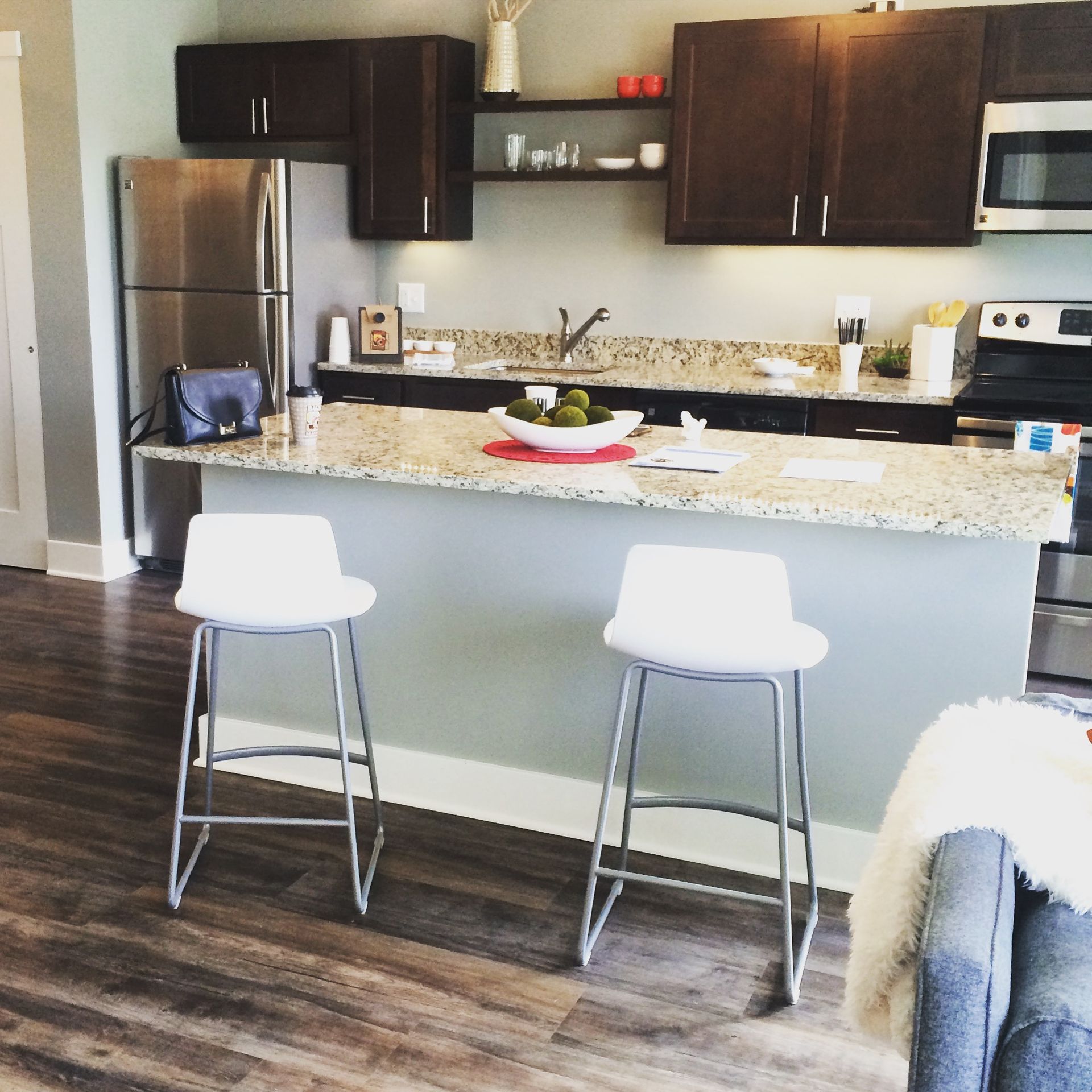 Modern kitchen with granite island, two white stools, dark cabinets, and stainless steel appliances