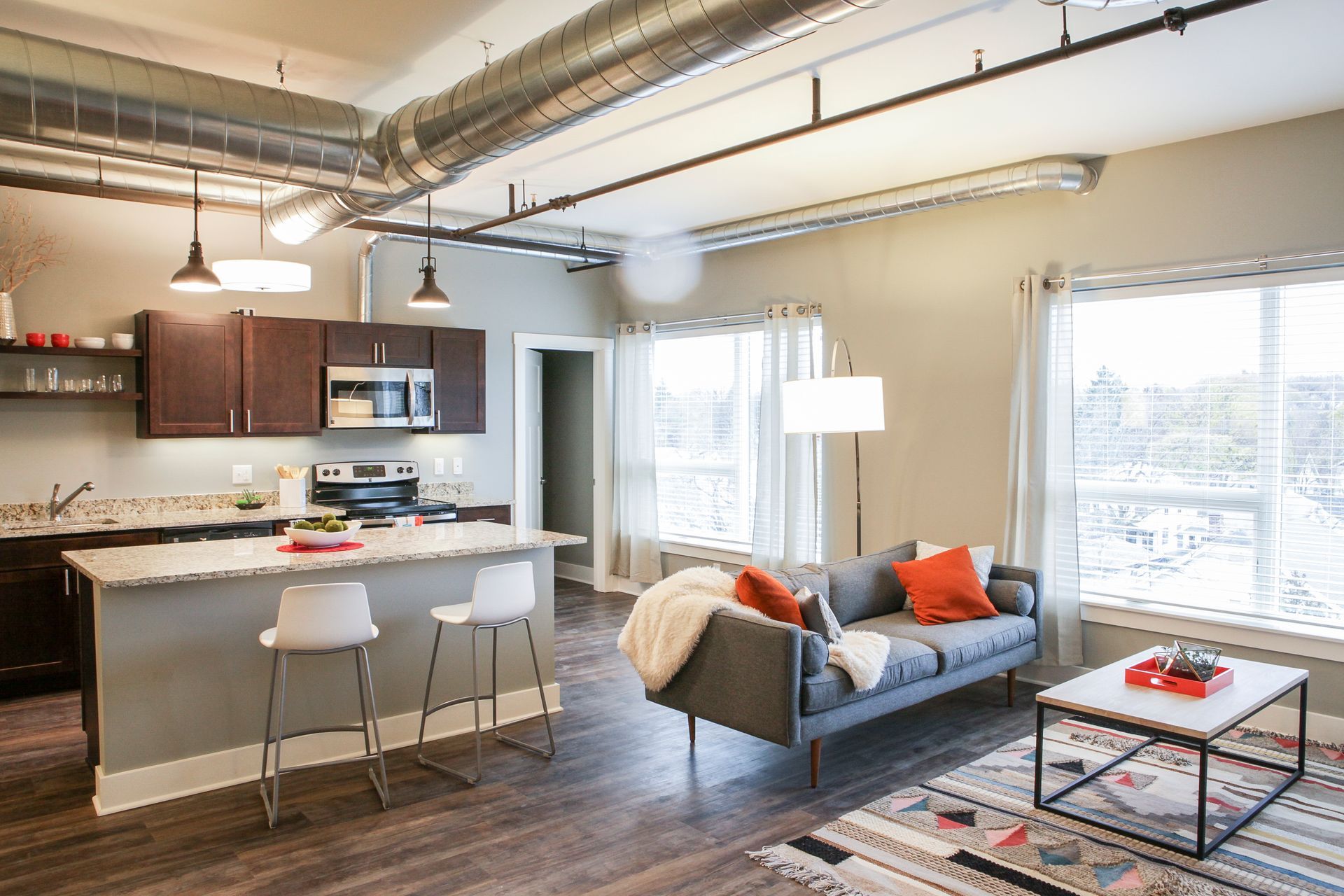 Bright modern loft living room with kitchen island, gray sofa, large windows, and exposed ceiling pipes