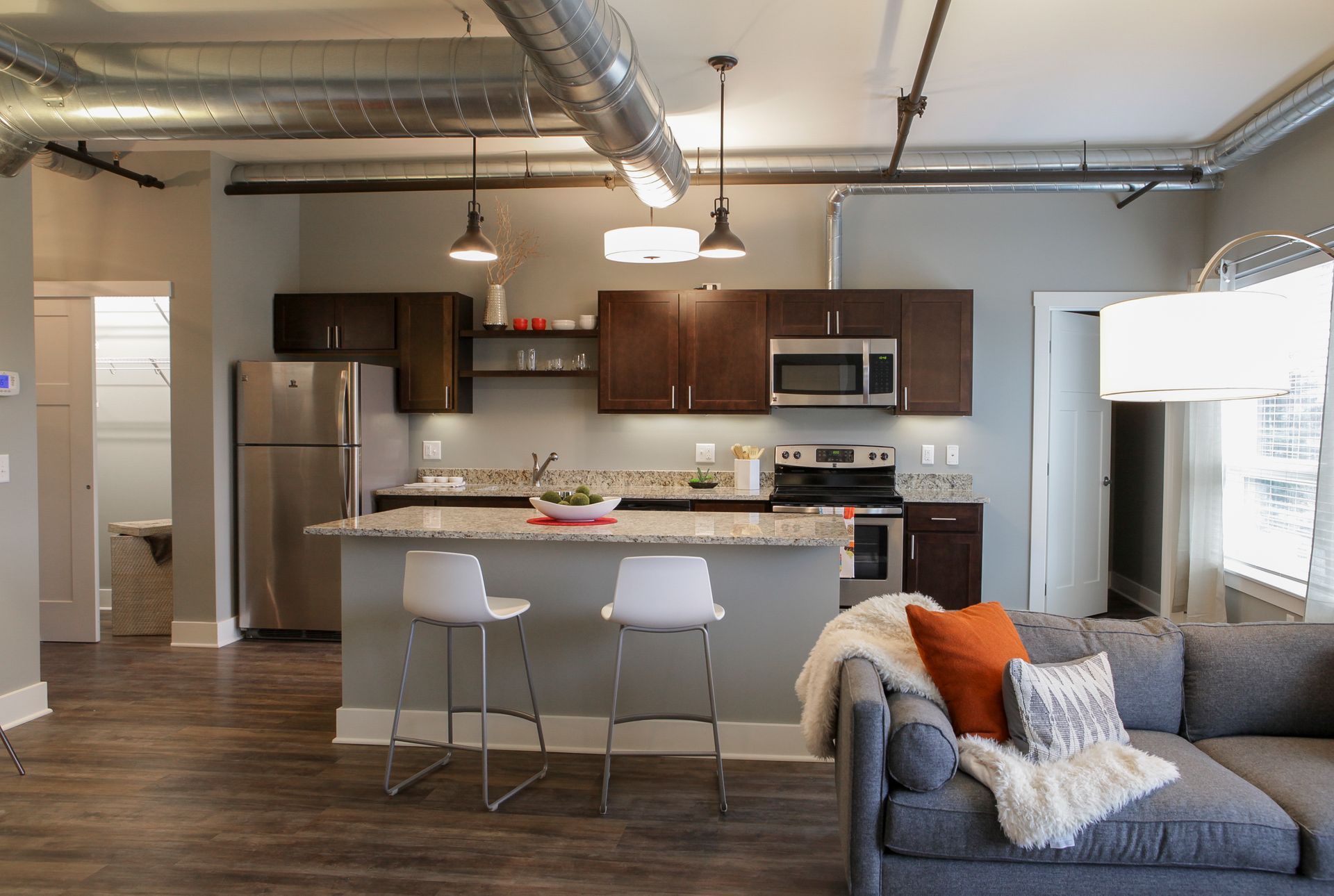 Open-concept kitchen and living room with stainless appliances, gray sofa, and industrial-style ceiling fixtures.