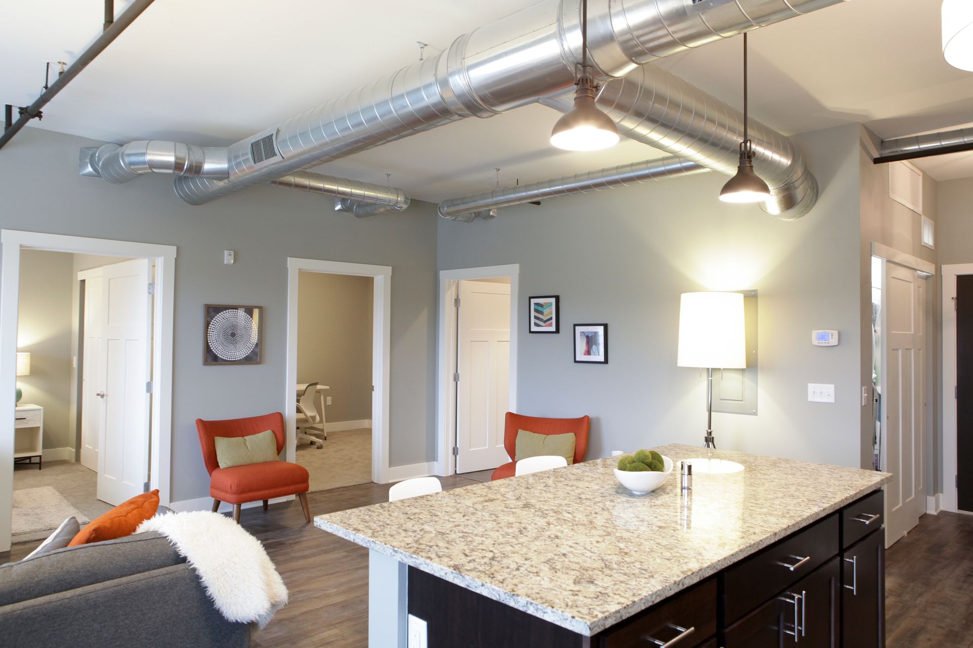 Modern open-concept living room with kitchen island, gray walls, exposed ductwork, and two orange chairs