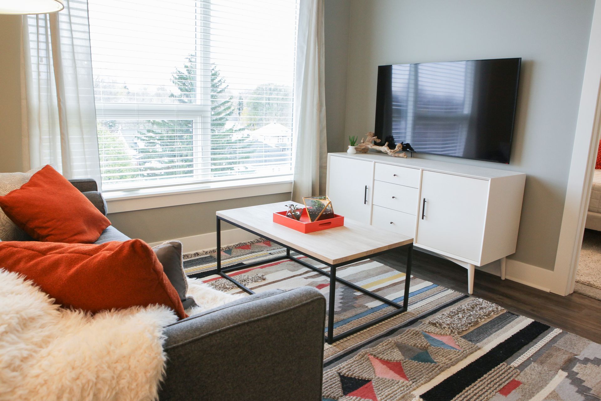 Bright living room with gray sofa, orange pillows, coffee table, TV, and large window with curtains