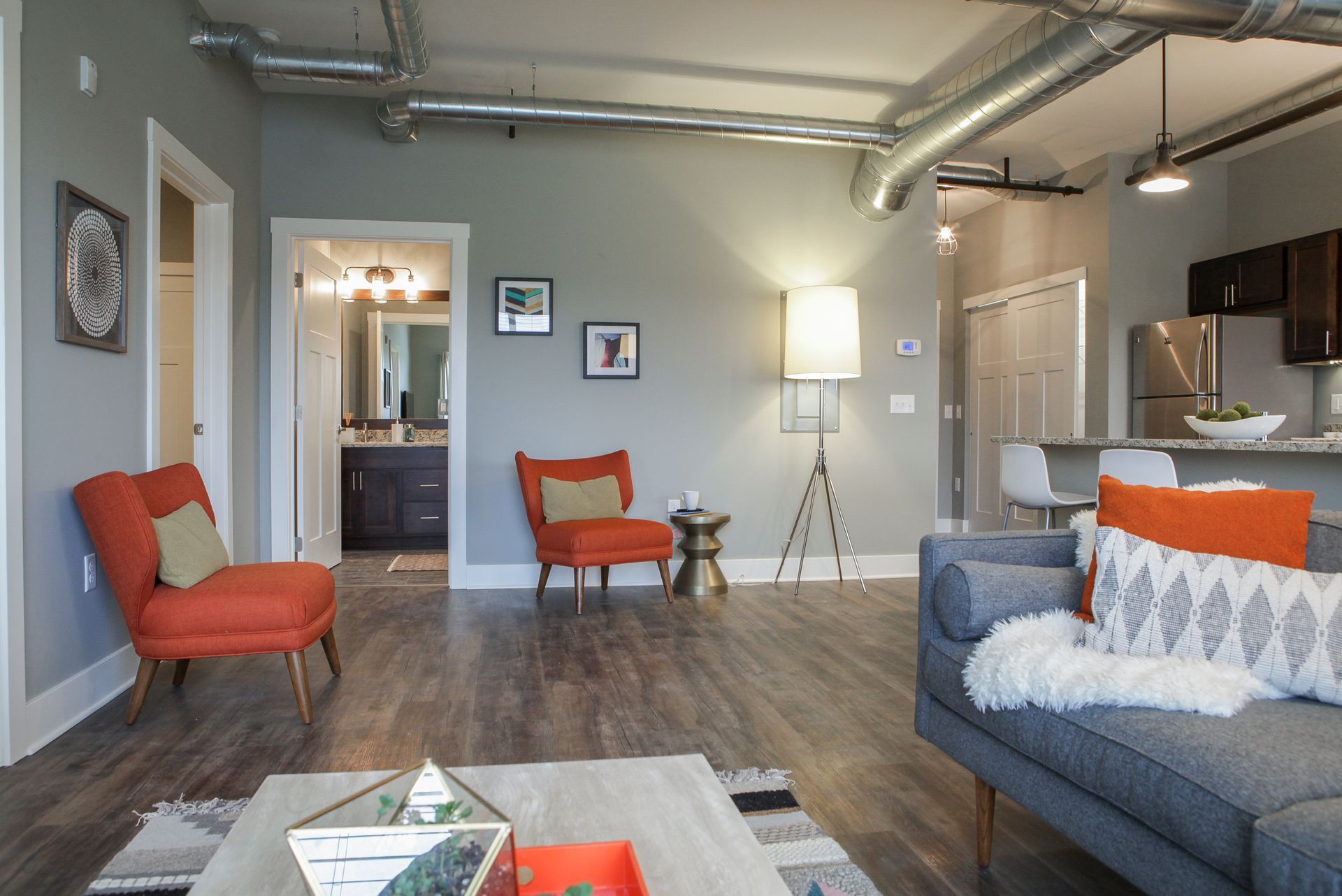 Modern living room with gray sectional, orange chairs, floor lamp, and open kitchen view
