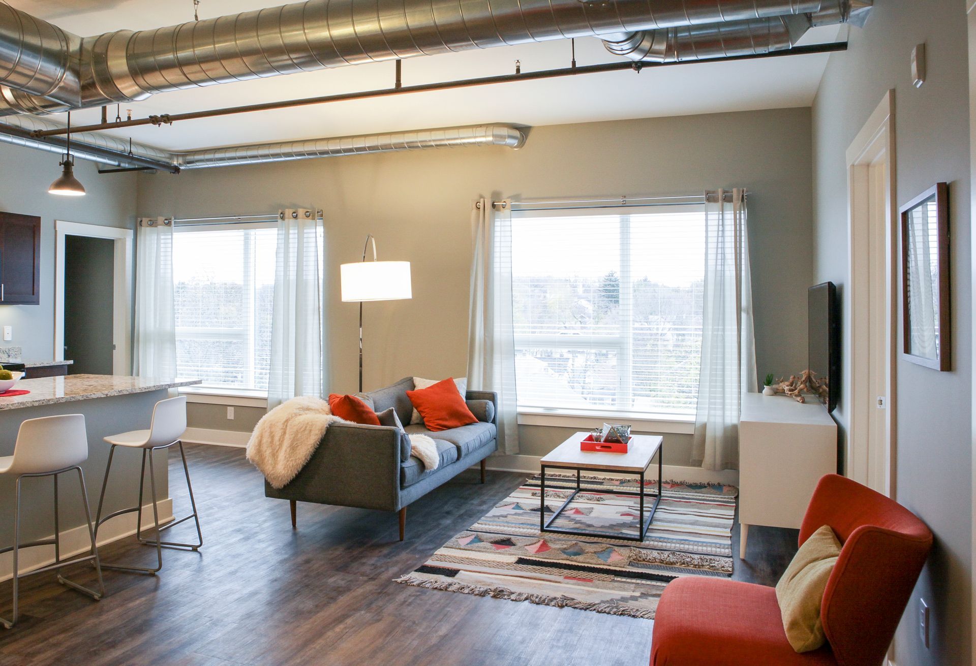 Modern living room with gray sofa, red chairs, large windows, exposed ceiling pipes, and colorful rug