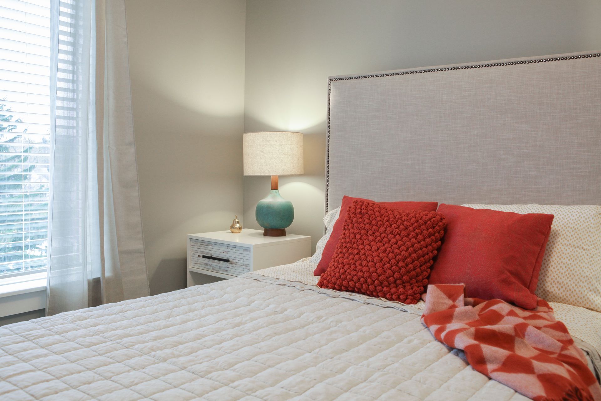 Bright bedroom with white quilt, red pillows, bedside lamp, and window with sheer curtains