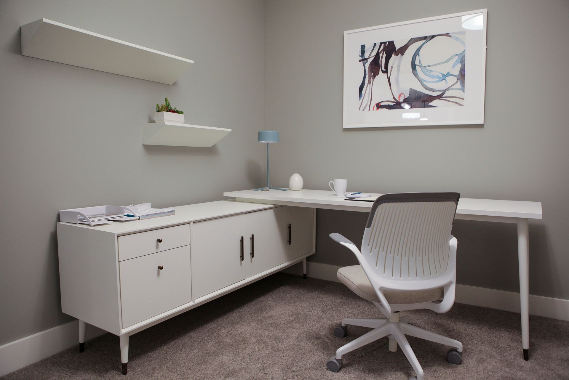 Modern home office with white desk, swivel chair, cabinet, shelves, and abstract wall art