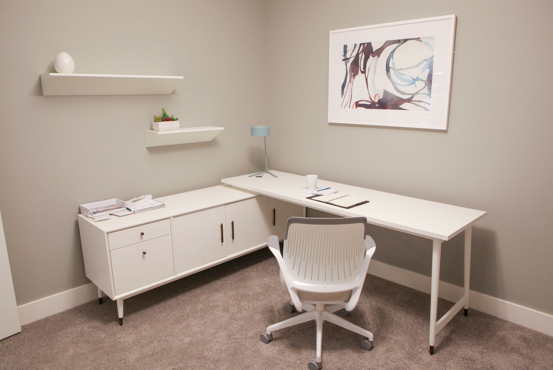 Minimal home office with white desk, chair, cabinet, shelves, and abstract wall art.