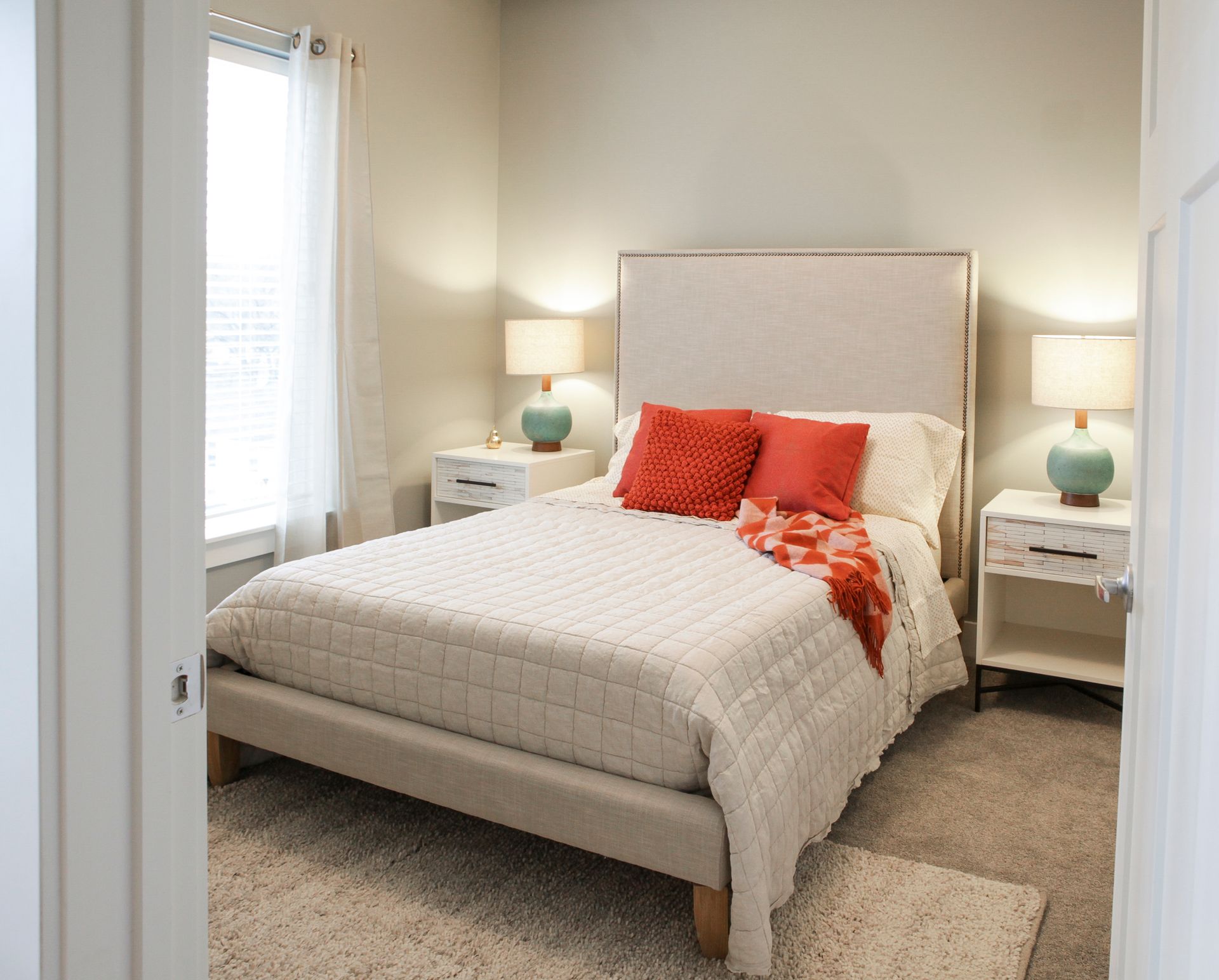 Bright bedroom with a neatly made bed, coral pillows, bedside lamps, and a window with sheer curtains