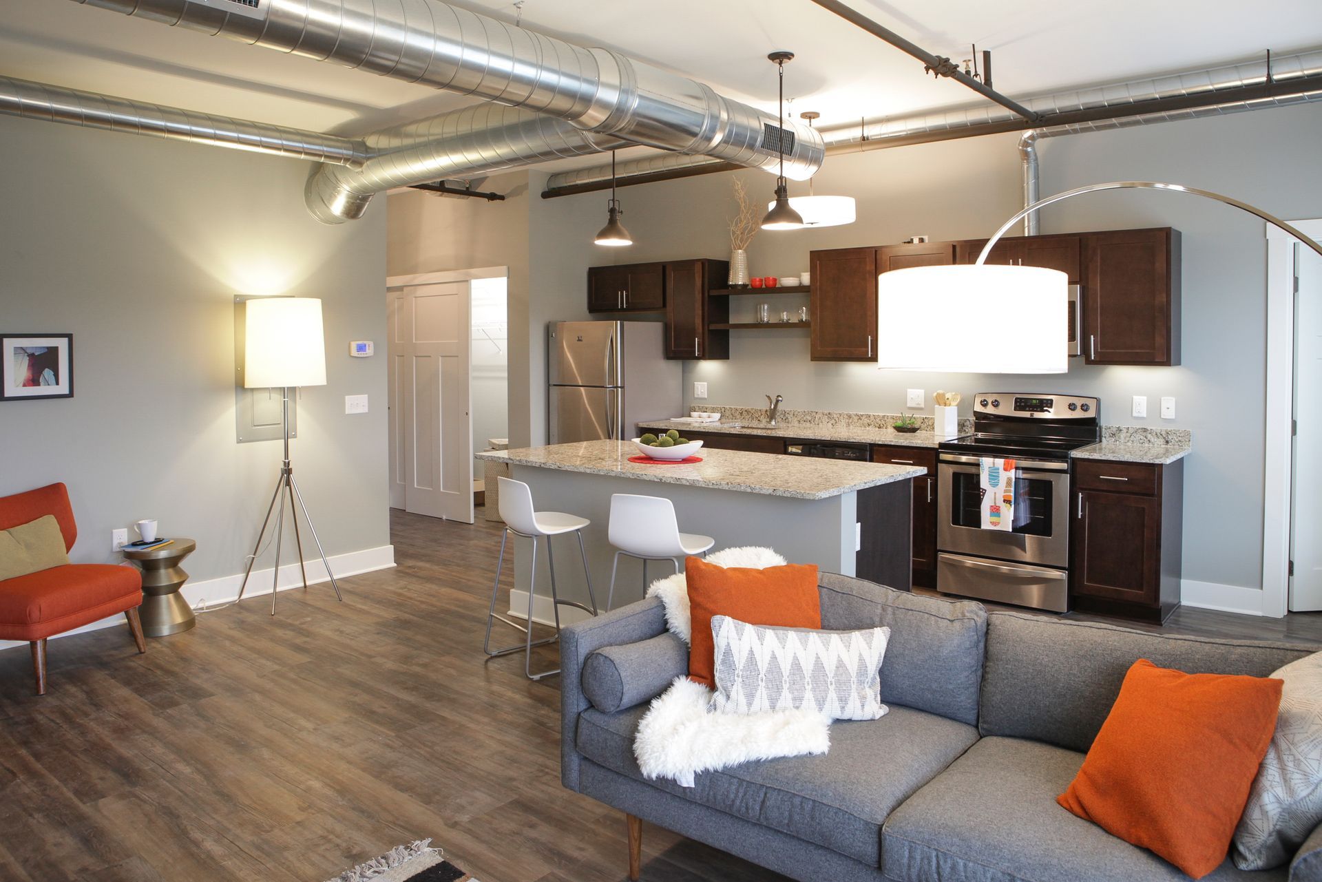 Modern open-plan living room and kitchen with gray sofa, orange accents, hardwood floors, and pendant lights