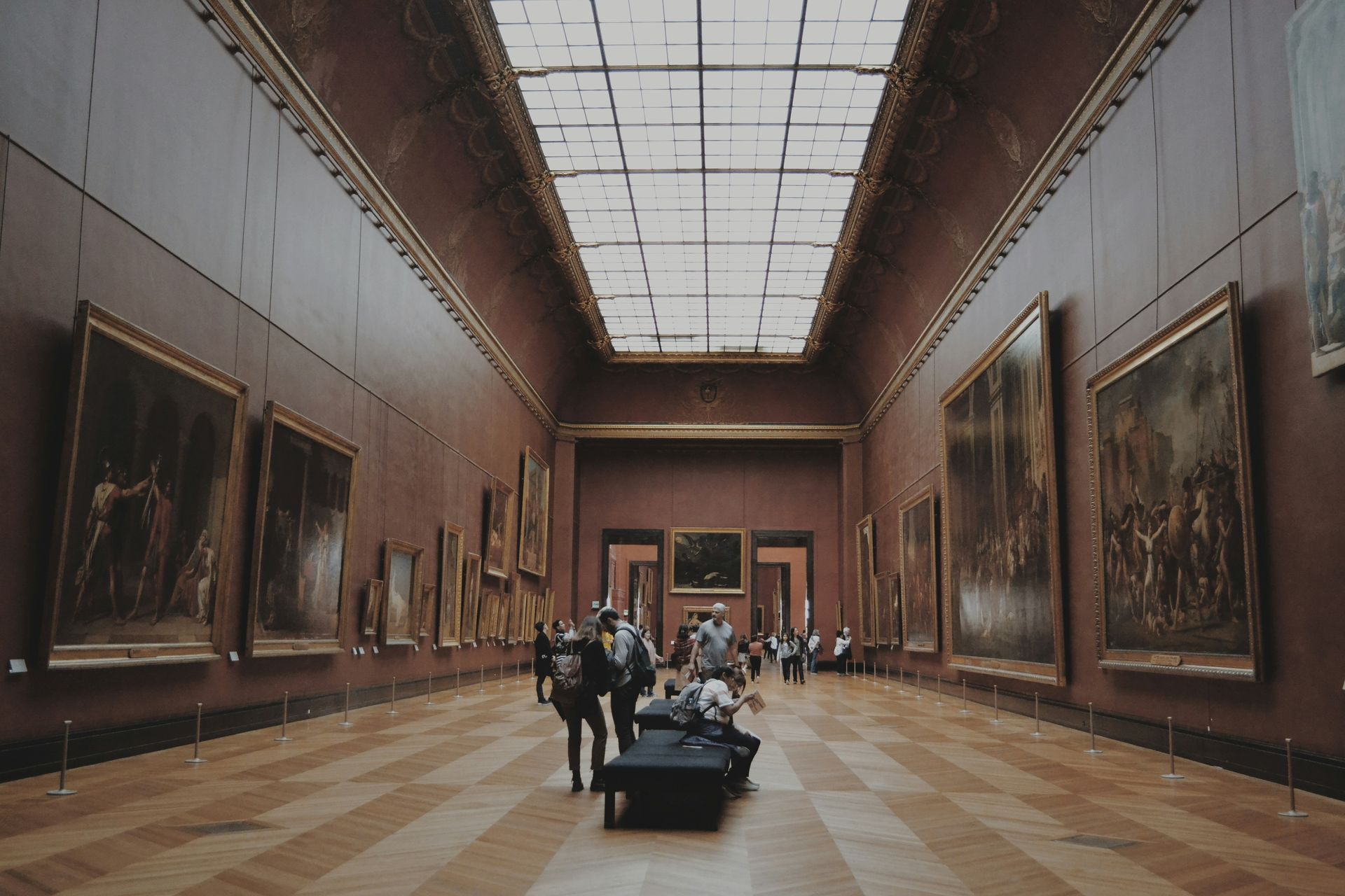 A grand art museum gallery with tall paintings, a skylight ceiling, and visitors walking through.