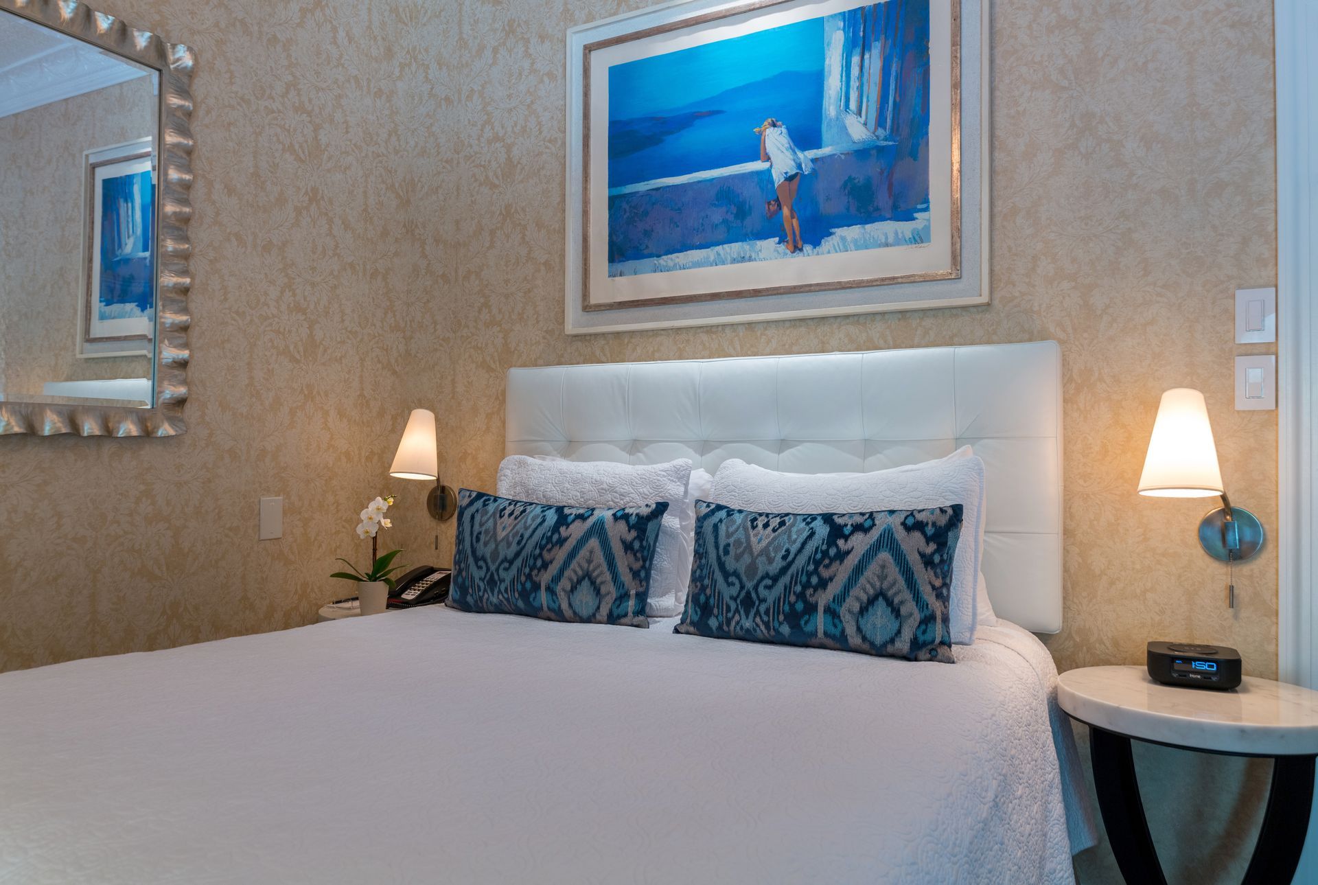 A hotel room with a bed , nightstand , mirror and painting on the wall.