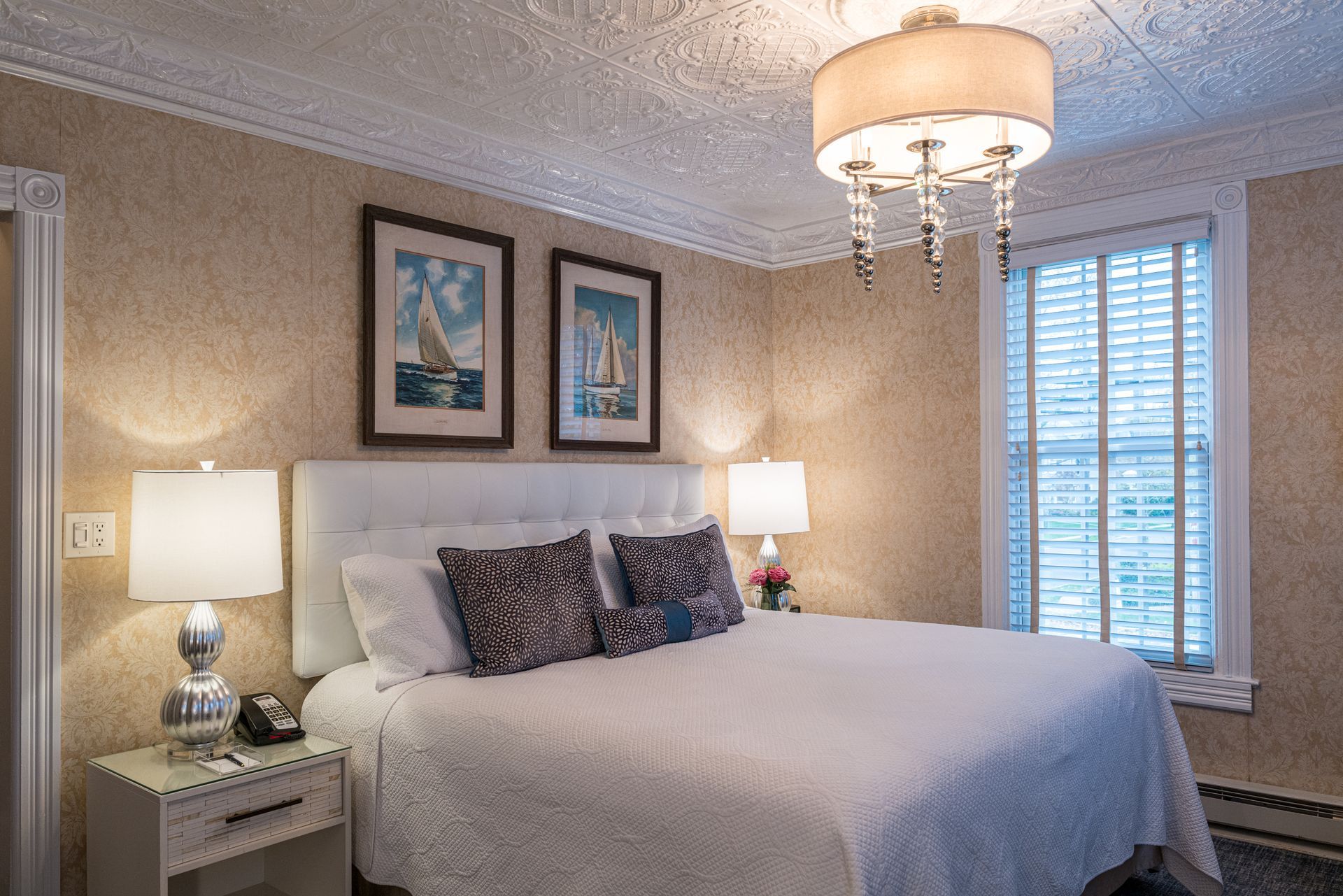 A bedroom with a king size bed , two lamps , and two sailboats on the wall.