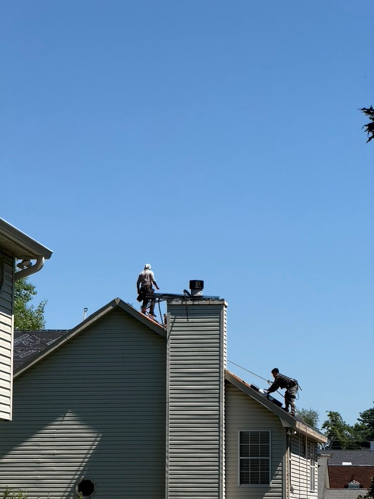 Workers on a house roof repairing shingles beside a brick chimney under a clear blue sky