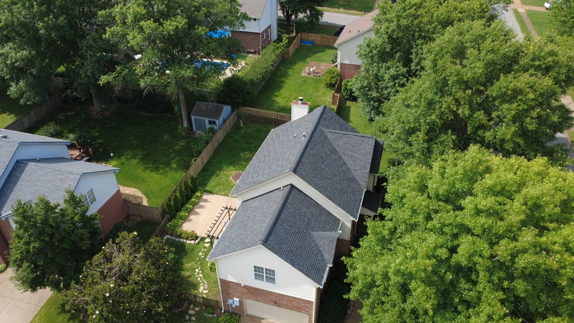 Aerial view of a suburban home with gray roof, trees, and neighboring houses on a sunny day