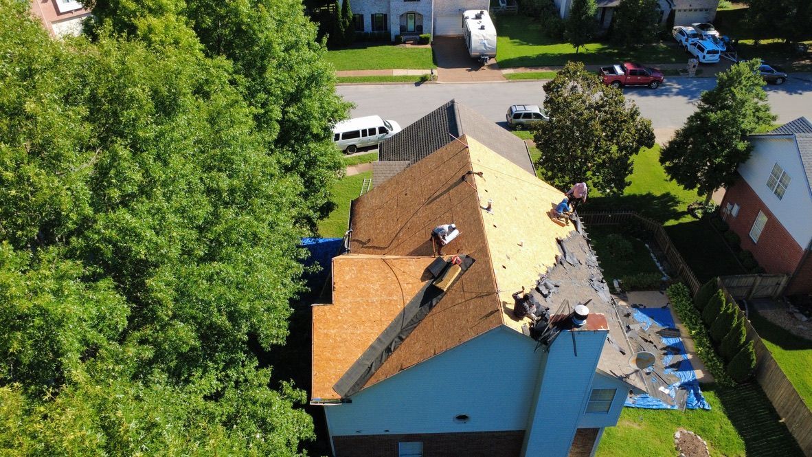 Aerial view of a house roof under repair, with workers and construction materials visible around the roof.