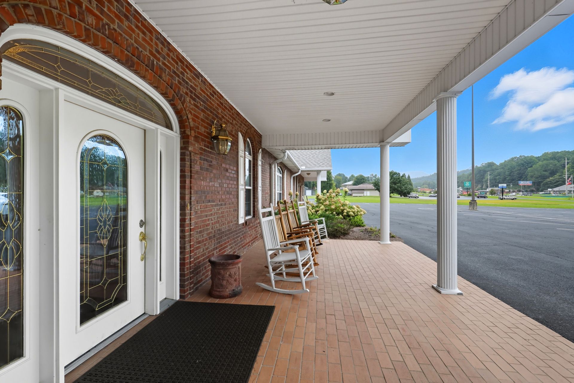 Brick storefront porch with white columns, double glass doors, rocking chairs, and a parking lot view