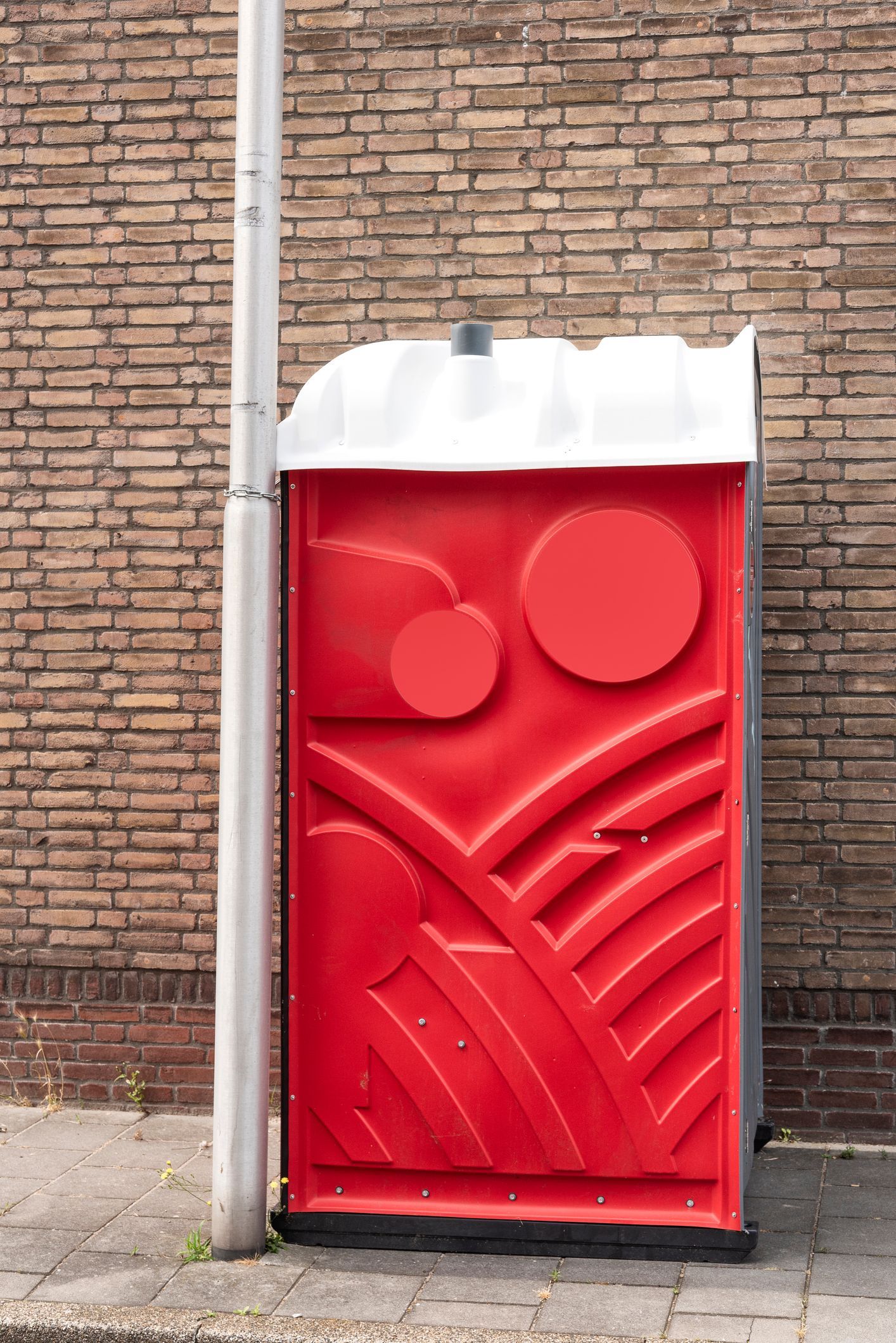 View of a Red Portable Toilet With a White Roof Against a Brick Wall