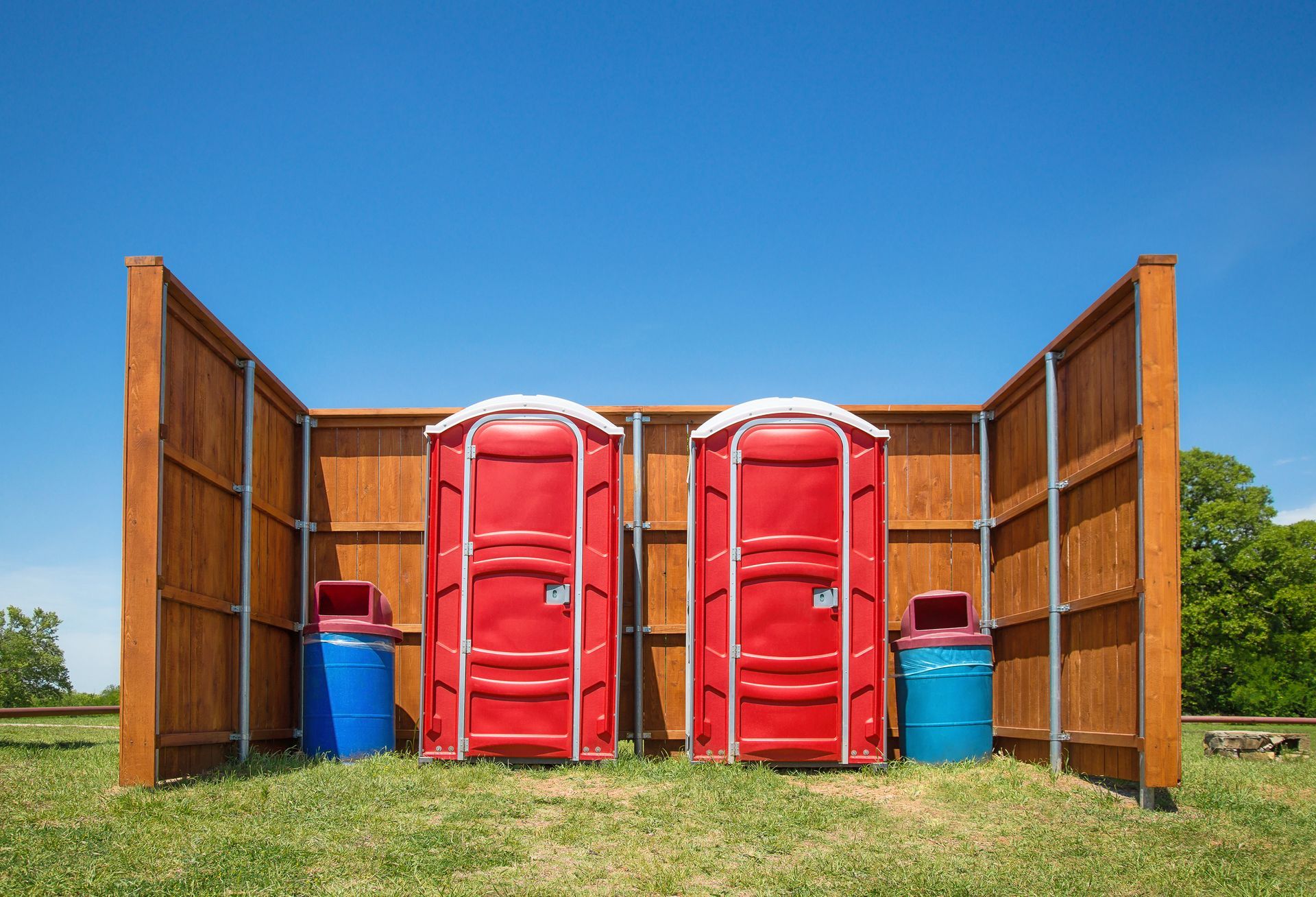 Two red portable toilets between wooden privacy screens, blue trash cans on grass, bright blue sky.