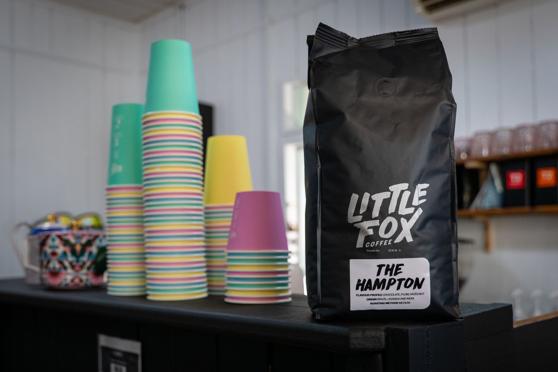 A black bag of Little Fox Coffee beans next to stacks of colorful paper cups on a counter.