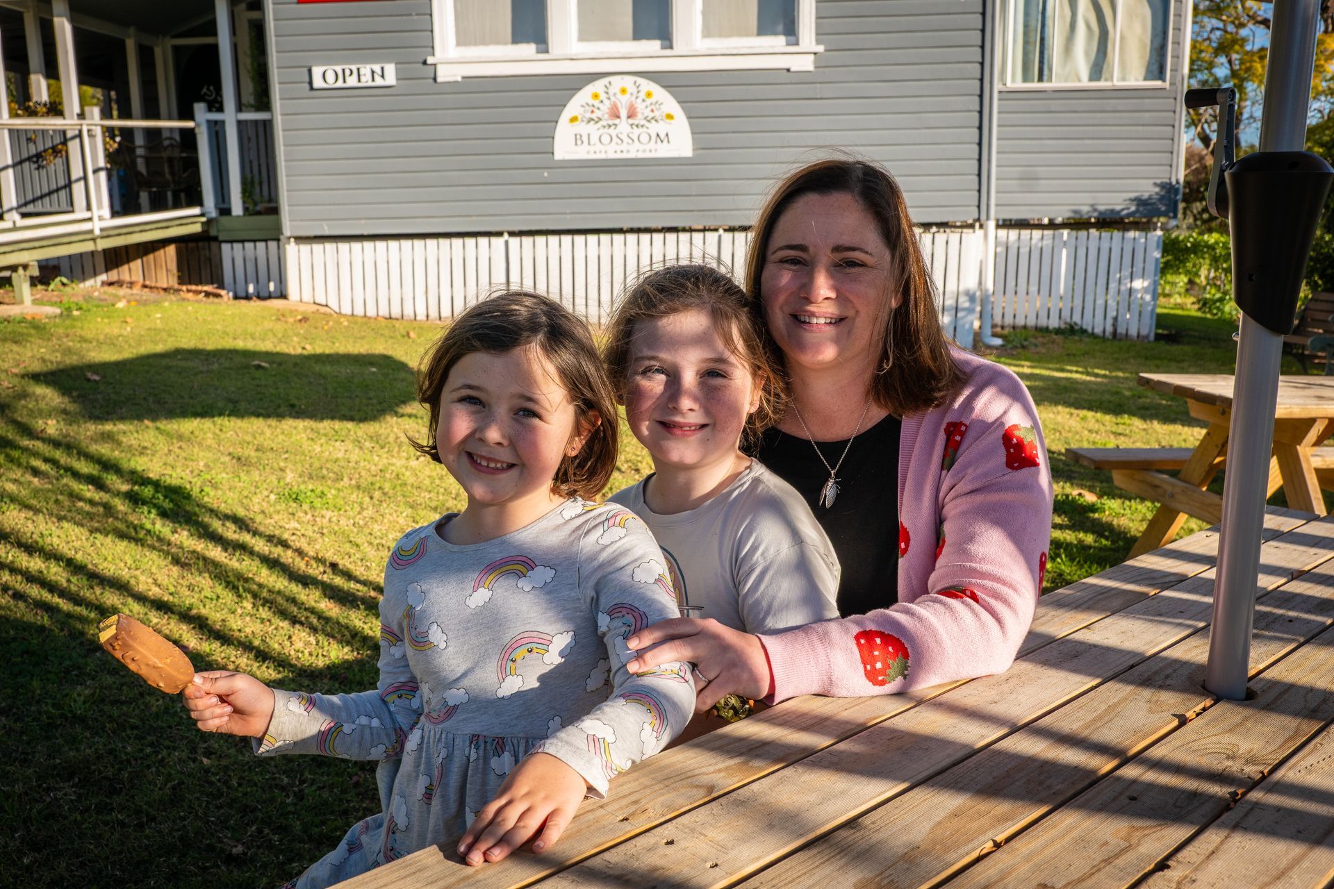 Woman and two girls smile at the camera, leaning on a picnic table outside a gray building.