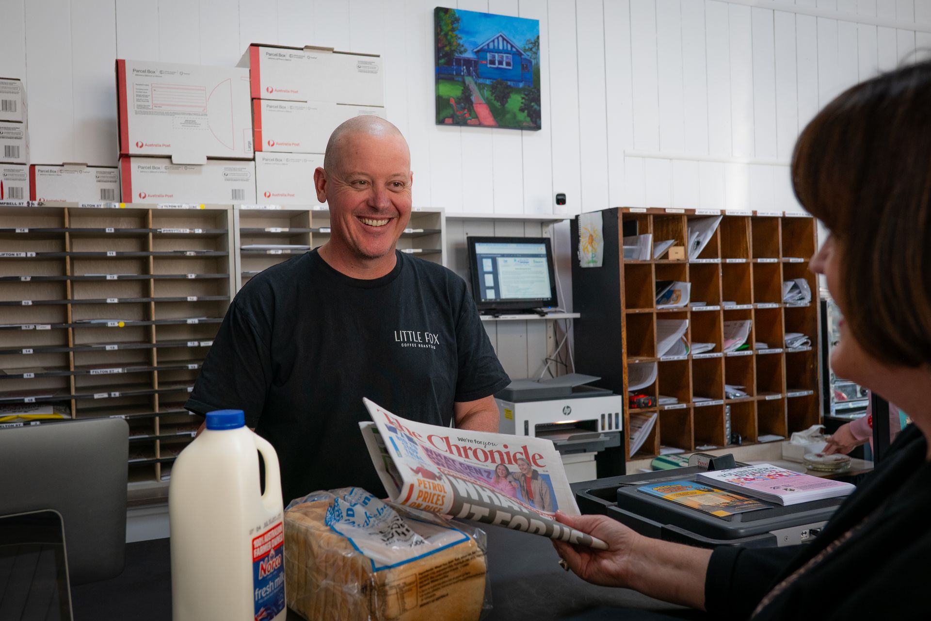 Man smiles at customer, handing over newspaper at counter in a shop. Milk and bread are visible.