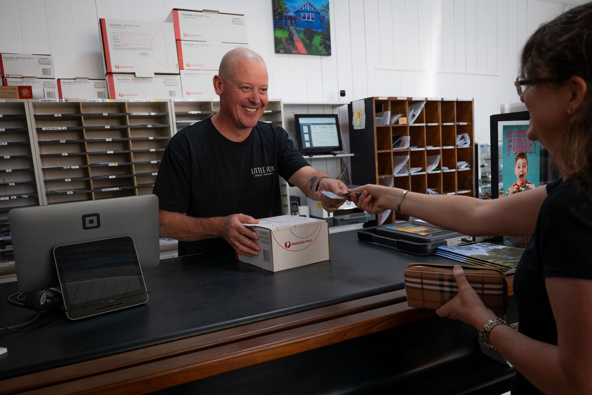 Man hands a package to a woman at a counter with a cash register; shelves in the background.