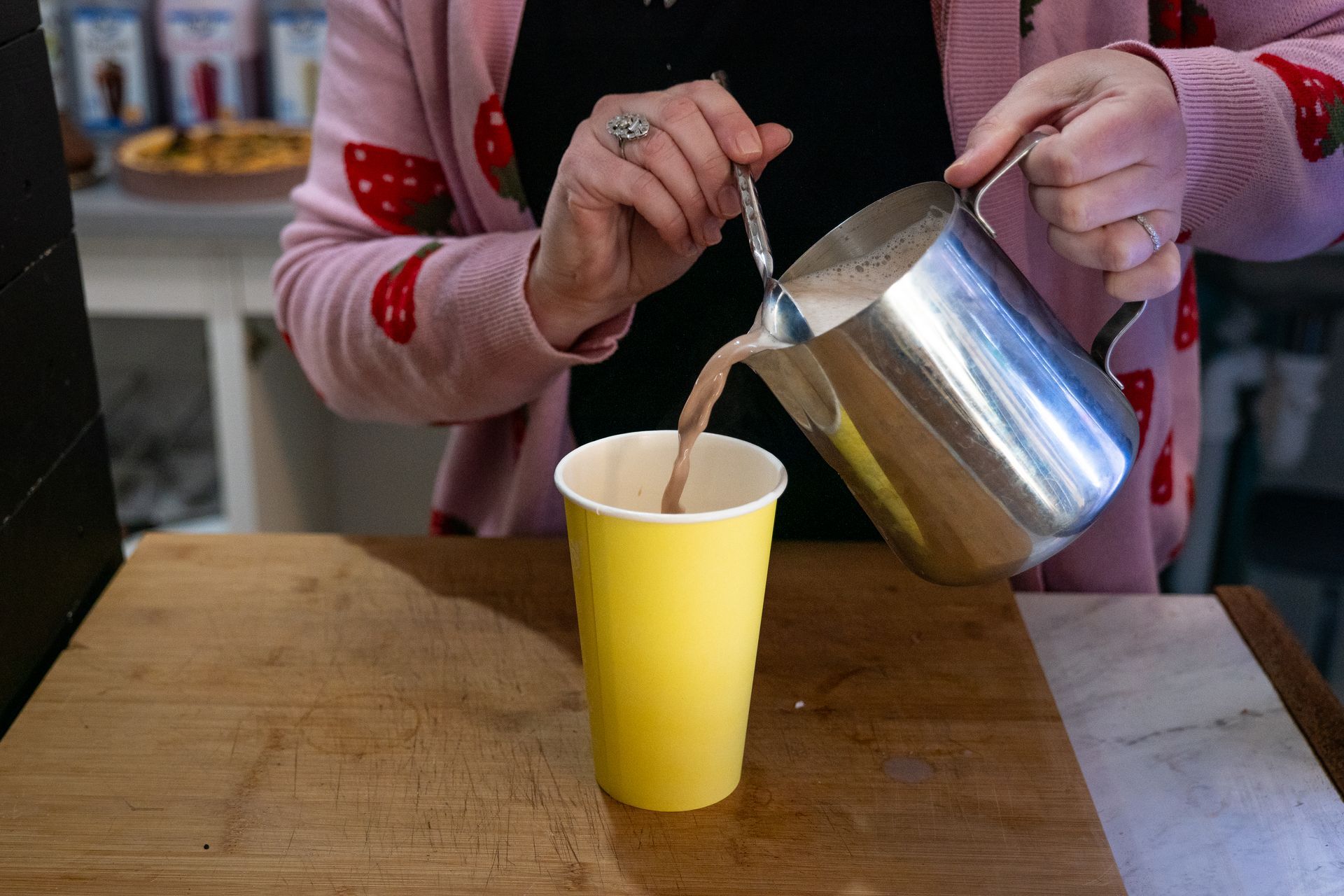 Person pouring chocolate drink from a metal pitcher into a yellow paper cup on a wooden surface.