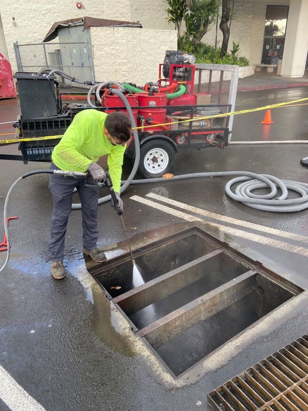 A person in a neon yellow shirt uses a high-pressure hose to clean an open storm drain in a parking lot.