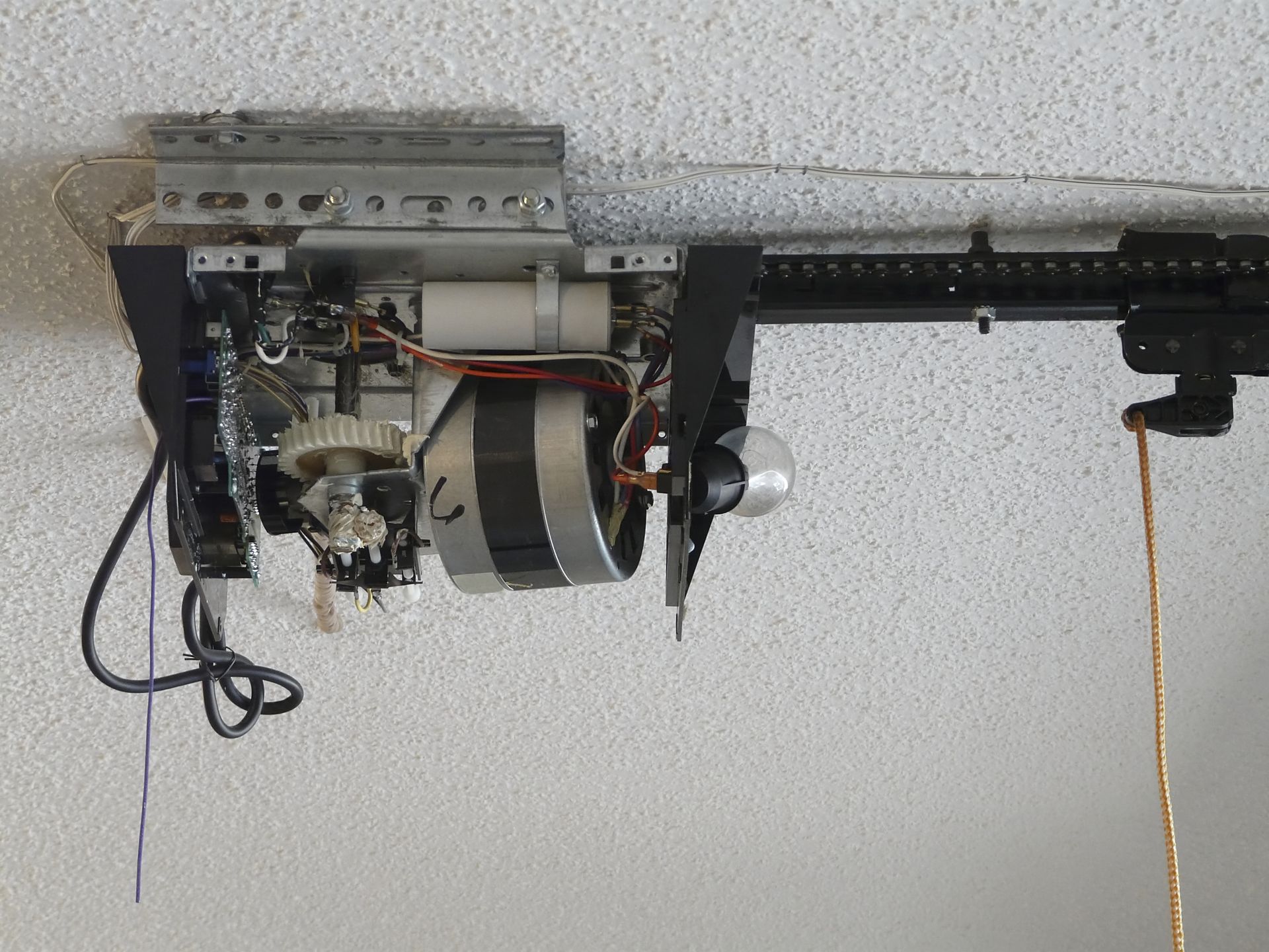 Opened garage door opener showing exposed motor, gears, wiring and drive rail on a ceiling