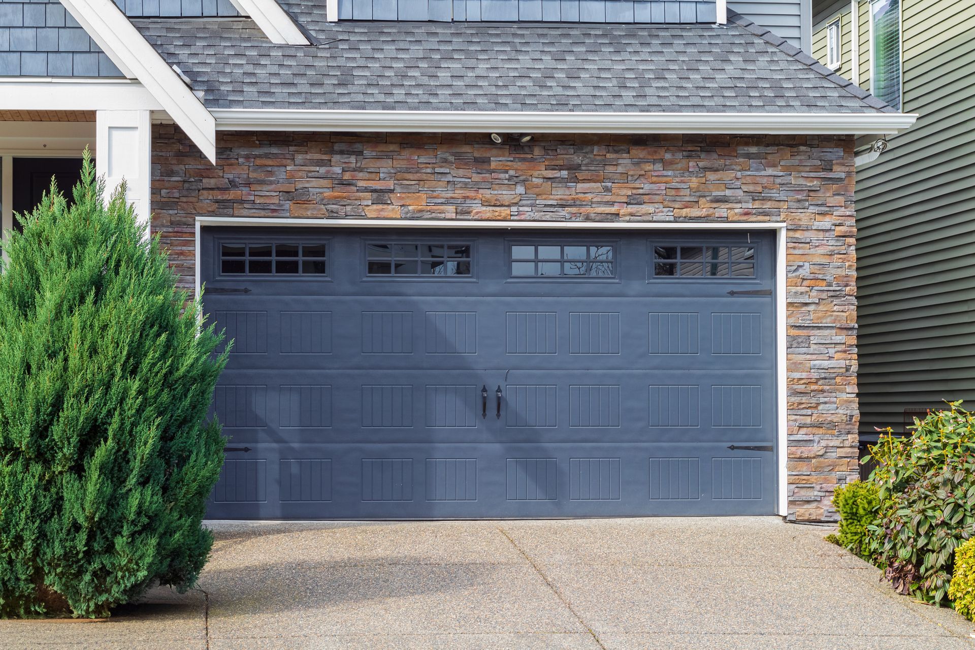 Blue garage door on a house with stone veneer; evergreen shrub in front.