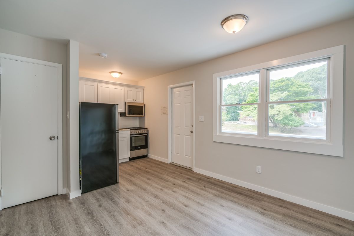 Empty apartment living area with kitchen, white walls, light wood floors, and two windows.