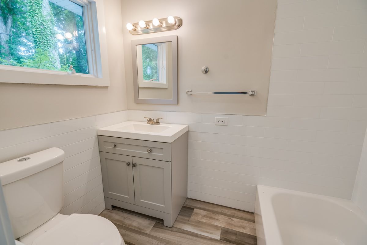 Bright bathroom with white tub, toilet, gray vanity, mirror, and window.
