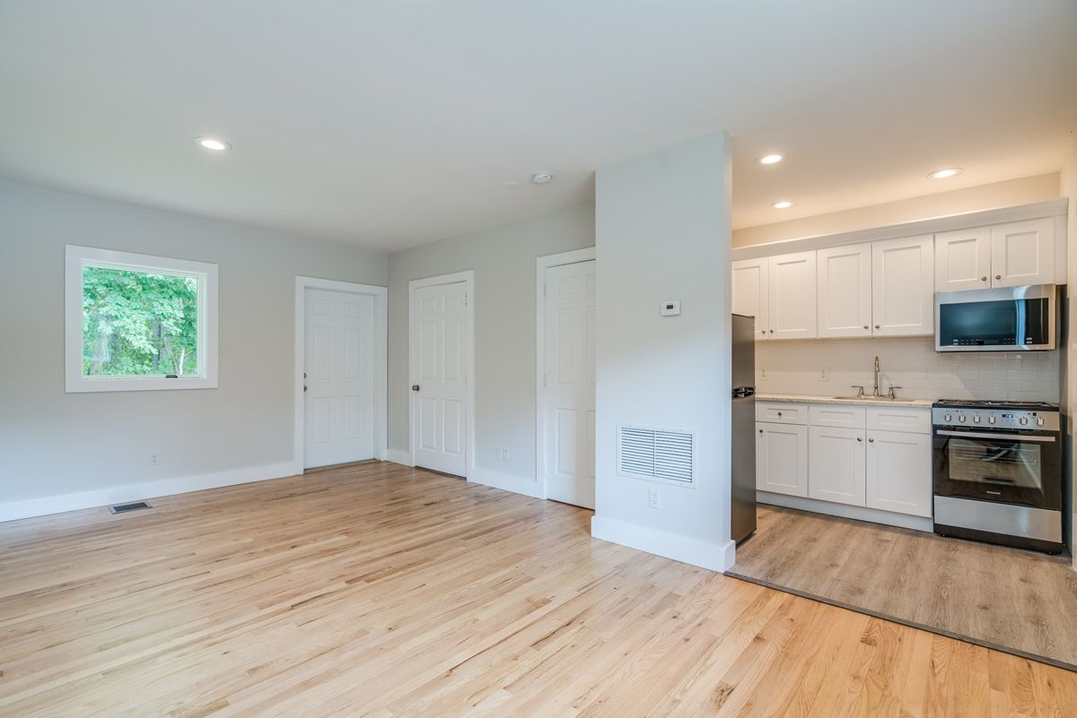 Empty apartment living room with light wood floors, white walls, and a small open kitchen.
