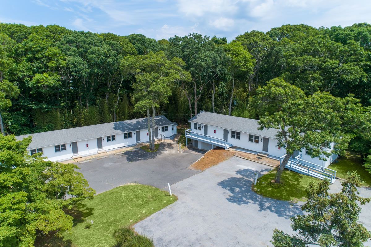 Aerial view of a long white building with a parking lot, surrounded by green trees.