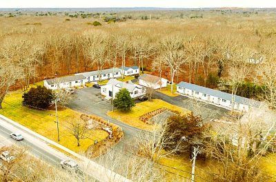 Aerial view of a white house and outbuildings beside a road, surrounded by leafless trees and grassy fields.