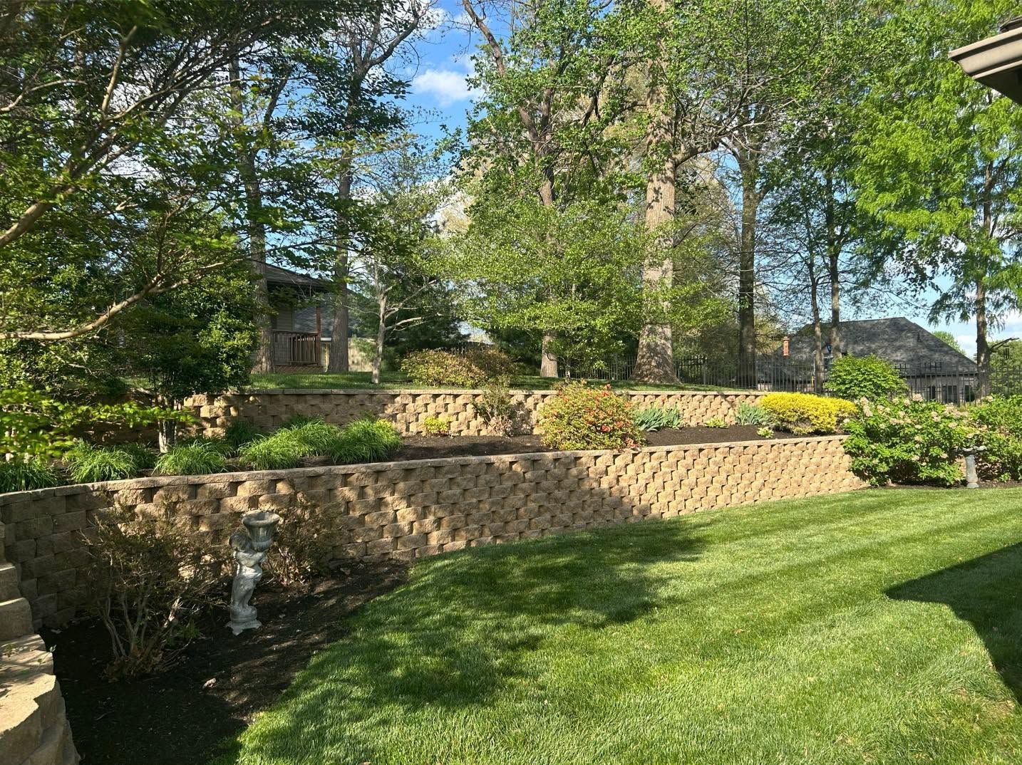 Terraced stone retaining walls and a green lawn beneath leafy trees in a sunny yard